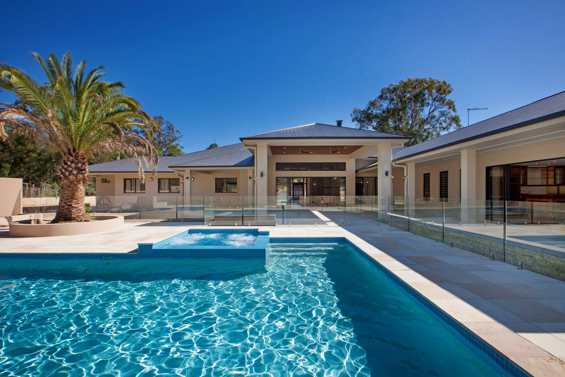 A modern backyard with a swimming pool, a small hot tub, a palm tree, and a contemporary house with large windows and a metal roof set against a clear blue sky.