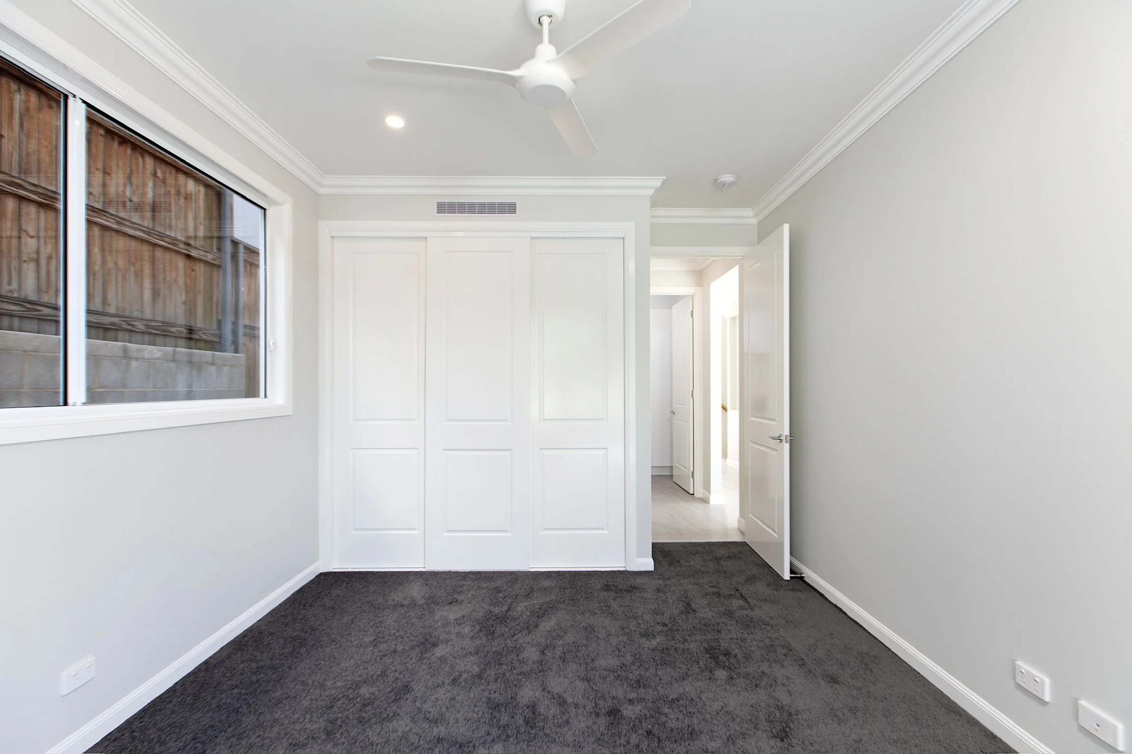 Empty room with white walls, a window with wooden blinds, dark carpet flooring, and an open door leading to a hallway.