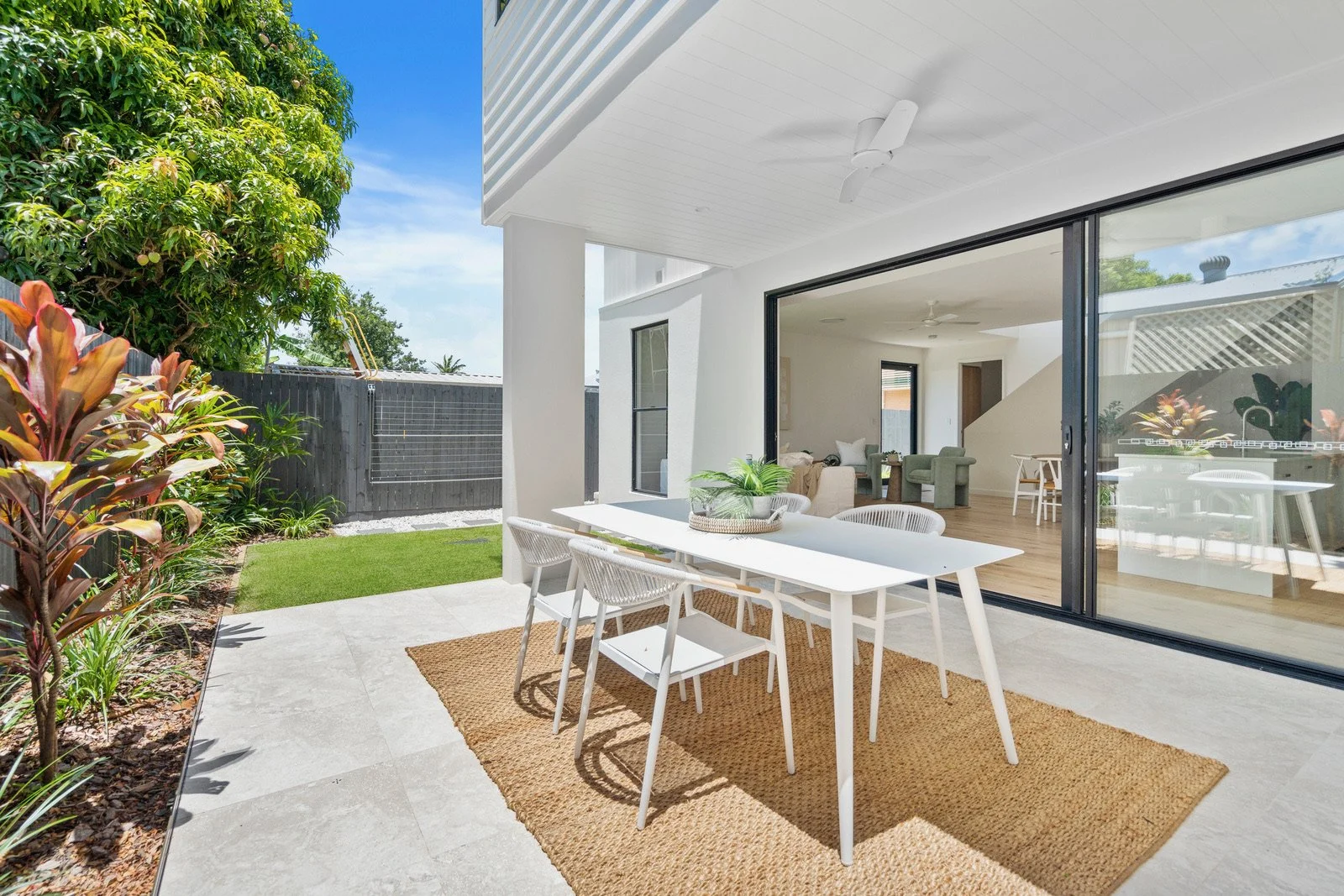 Outdoor patio with white dining table and chairs, green plants, and a view into a modern open living room with seating area and kitchen, surrounded by greenery and a dark wooden fence.