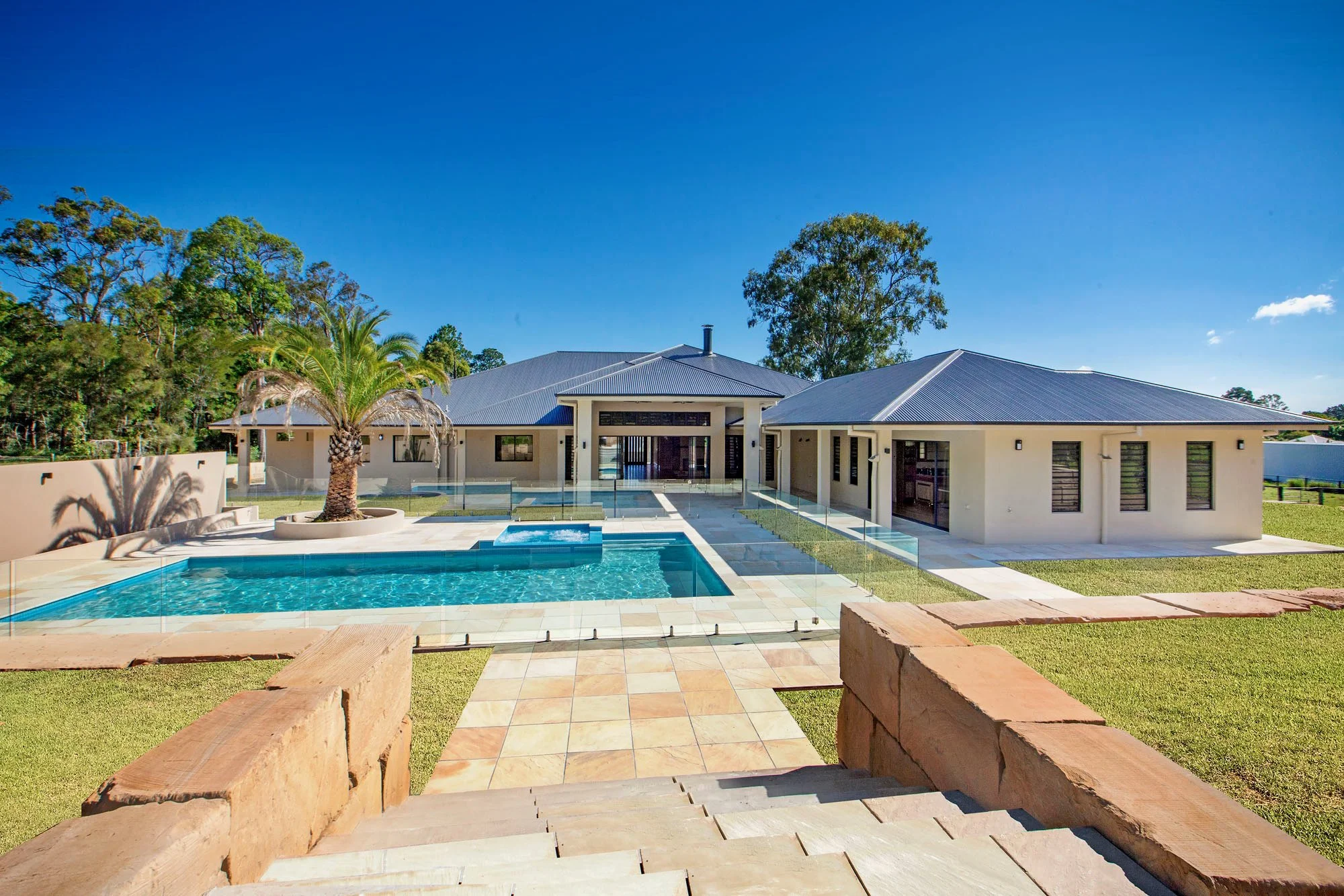 Modern house with a swimming pool in a sunny backyard, surrounded by green trees and a clear blue sky.