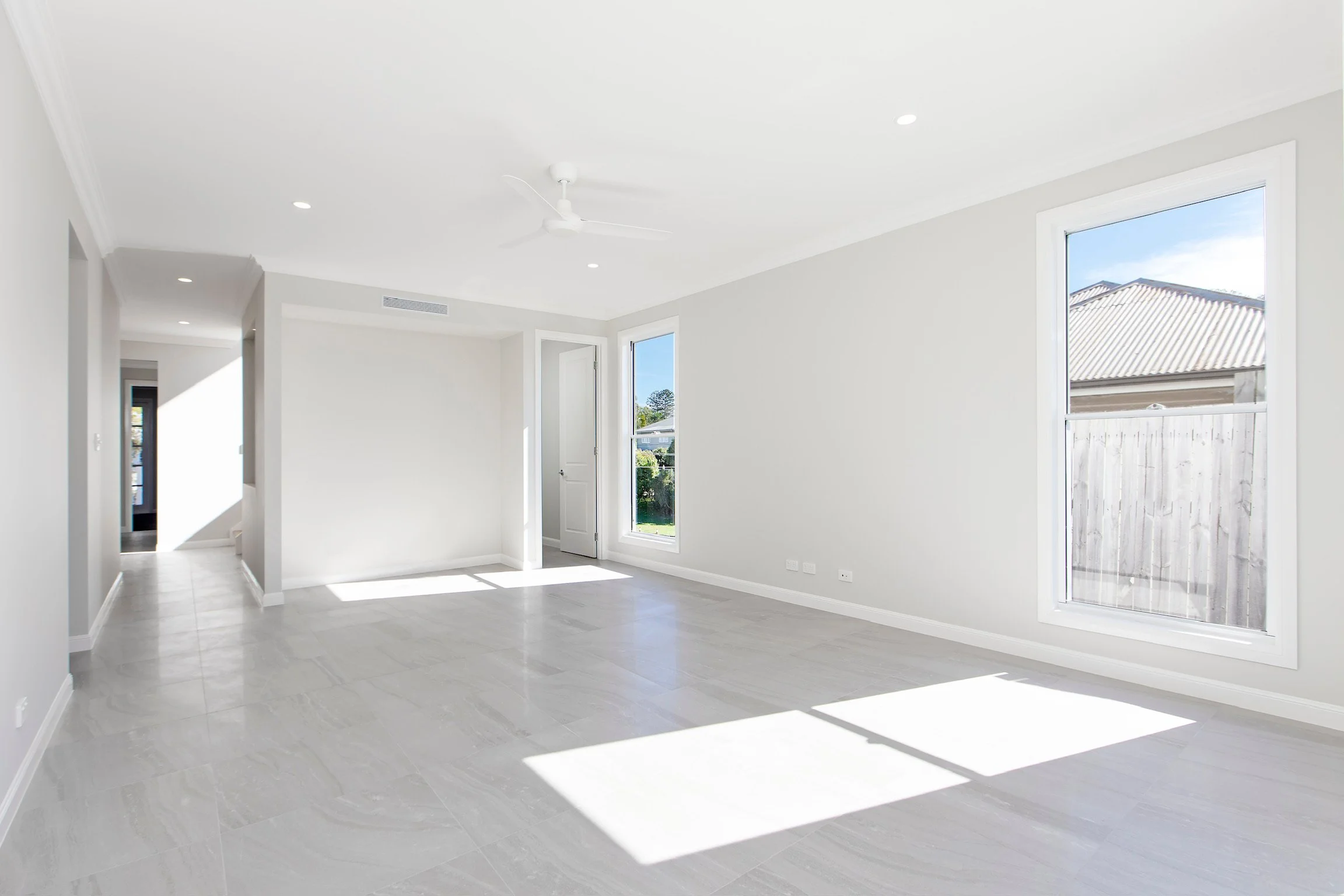 A spacious, empty living room with white walls and large windows letting in natural sunlight. The room has light-colored tiled floors, a white ceiling with a ceiling fan, and appears clean and modern.
