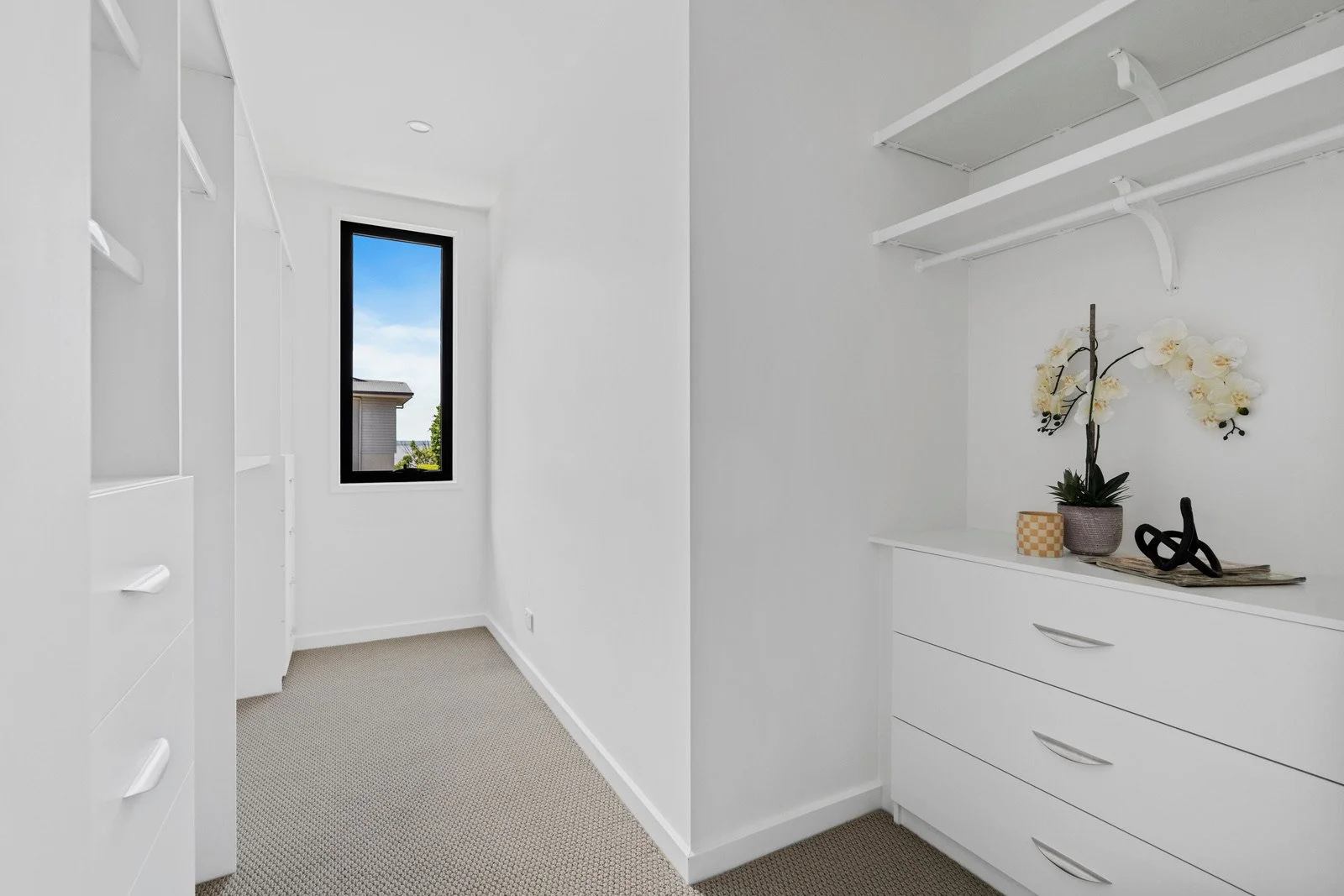 A small walk-in closet with white walls, white built-in shelves and drawers, and a window showing a house and trees outside. Decor items like plants and artwork are on the dresser.