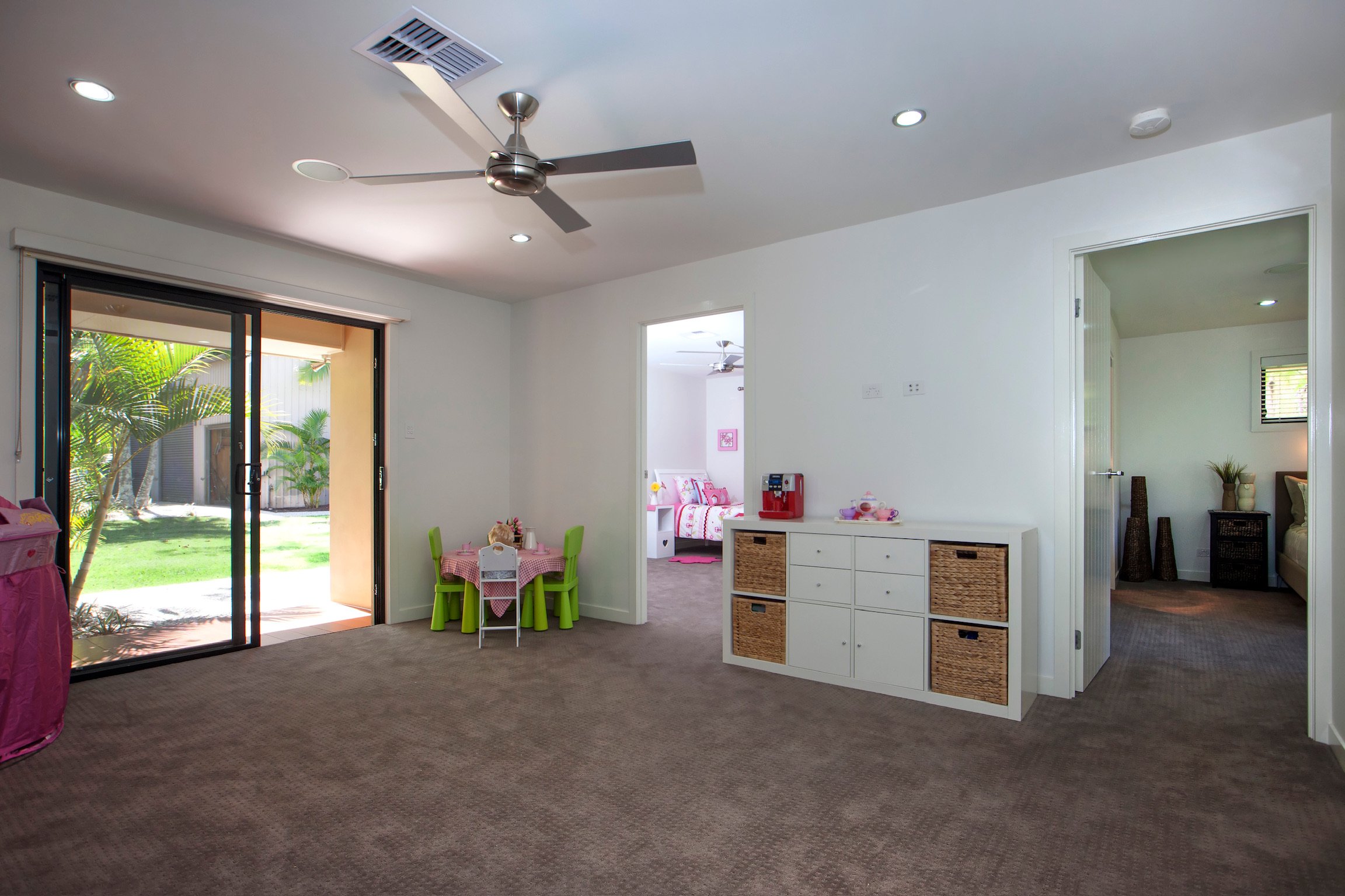 Living room with sliding glass door leading outside, small pink child's table with four chairs, white storage unit with baskets, and a doorway to a bedroom with pink bedding.