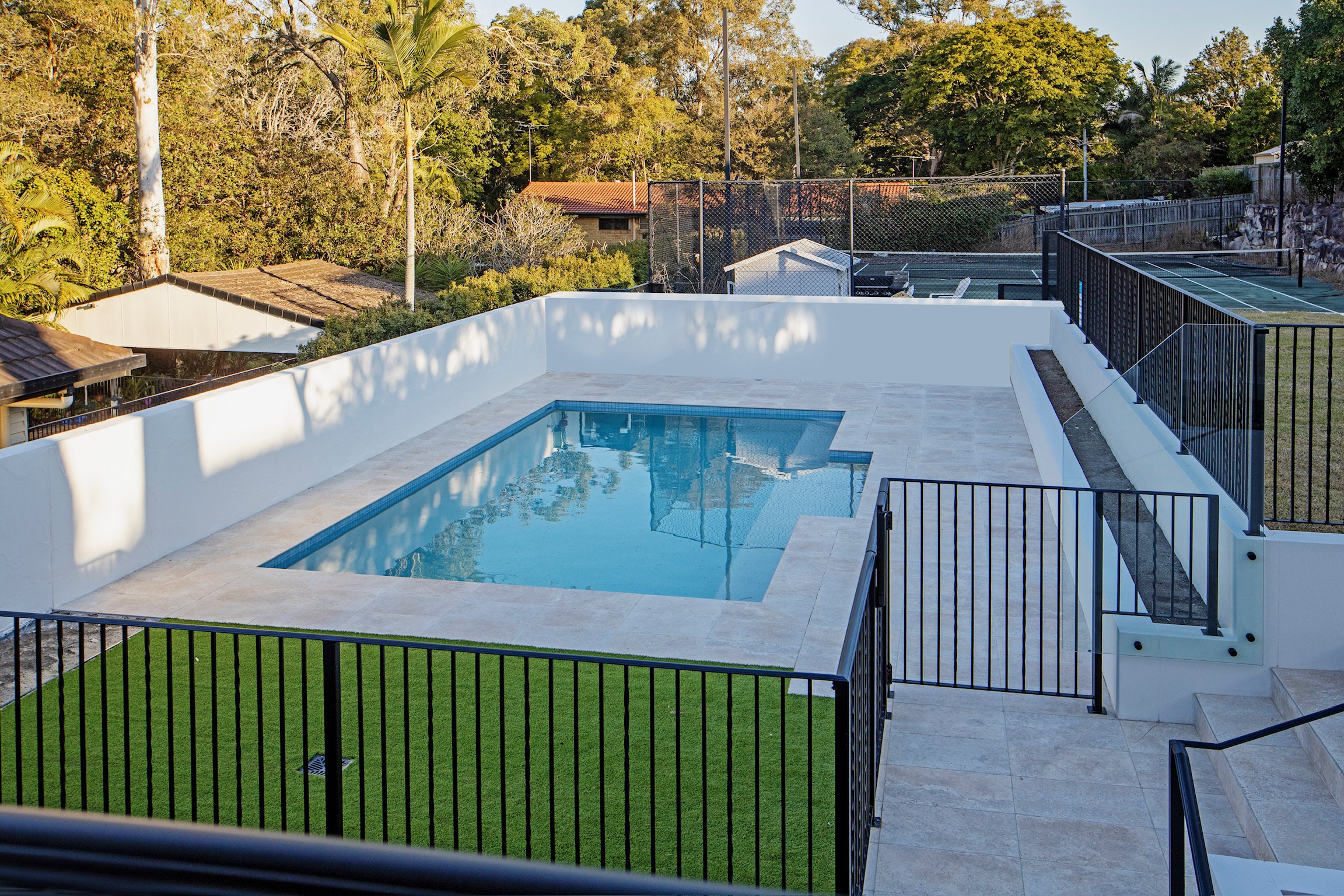 A backyard with a pool, surrounded by a white wall and black metal fencing, with a grassy yard area, trees, and tennis courts in the background.