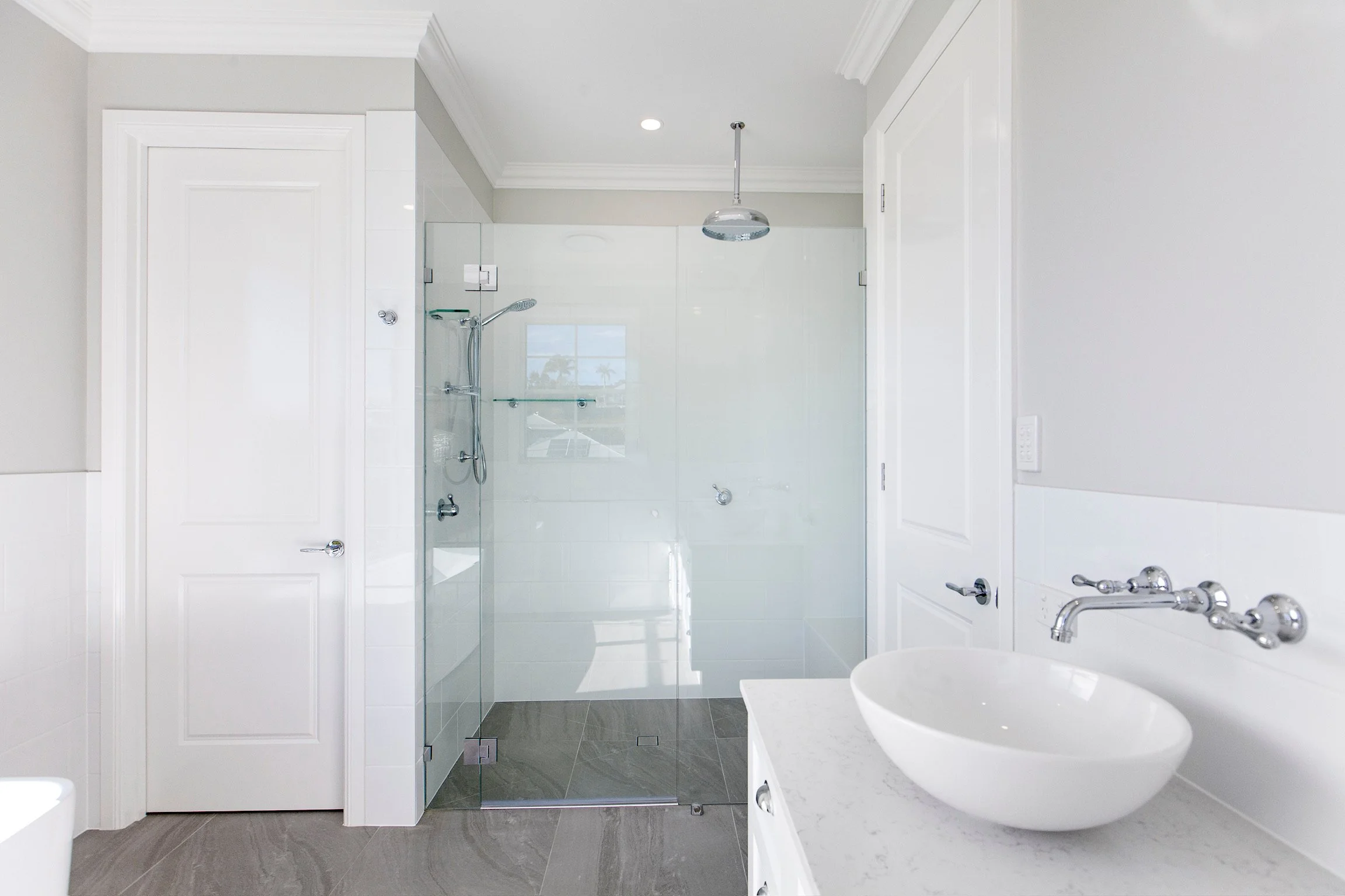 Modern bathroom with a glass-enclosed shower, white walls, a white door, a white sink with a chrome faucet, and gray tiled flooring.