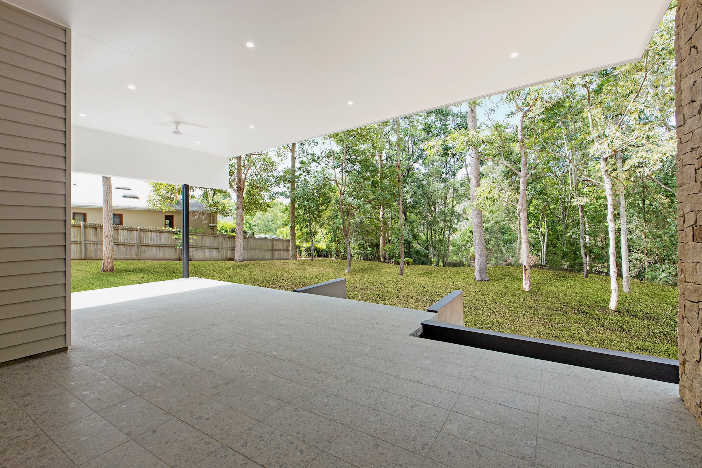 View of a covered outdoor patio with tiled flooring, overlooking a grassy backyard with trees and a wooden fence.