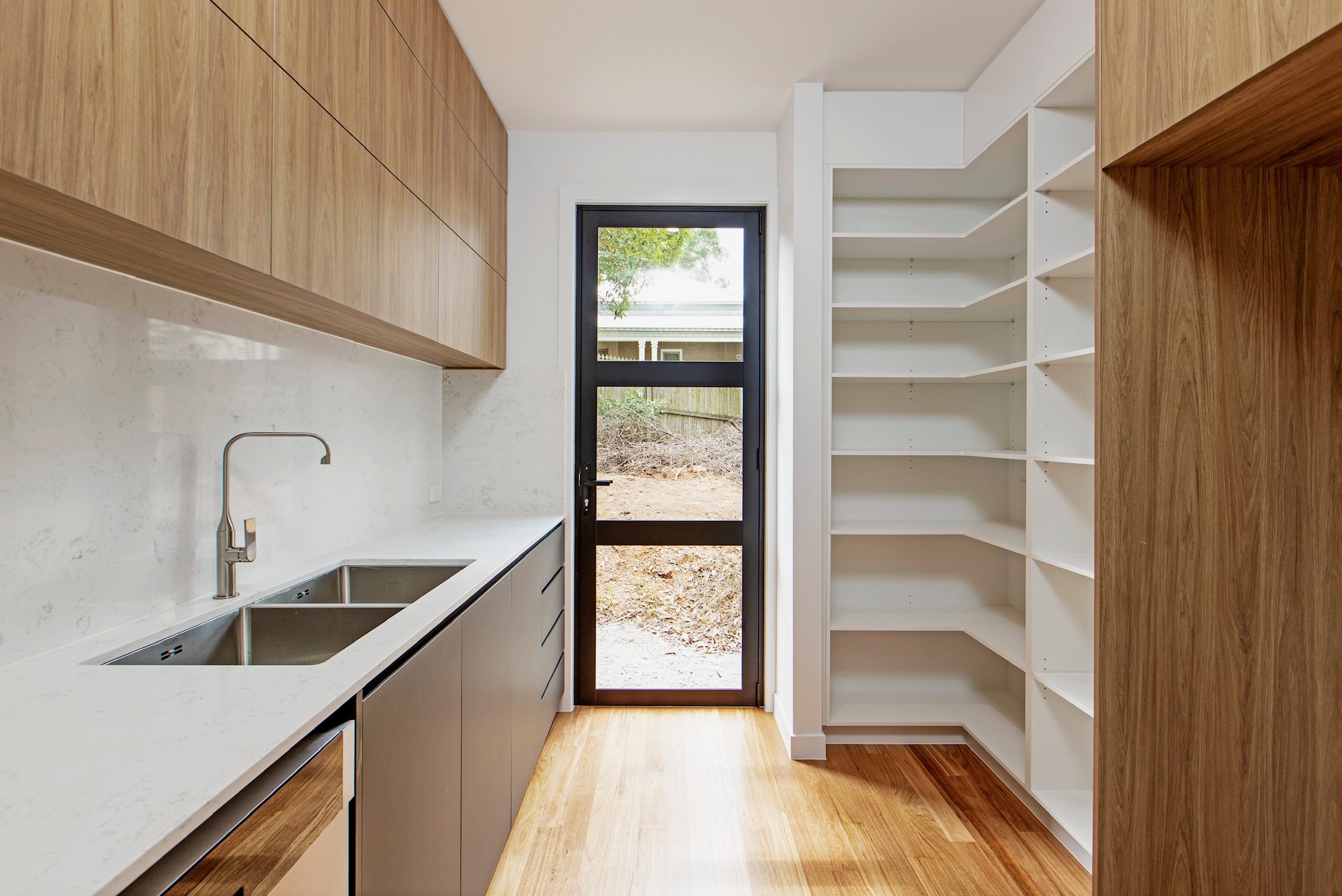 Modern kitchen with white countertops, wooden upper cabinets, and white open shelving, featuring a door leading outside with a view of a yard and trees.