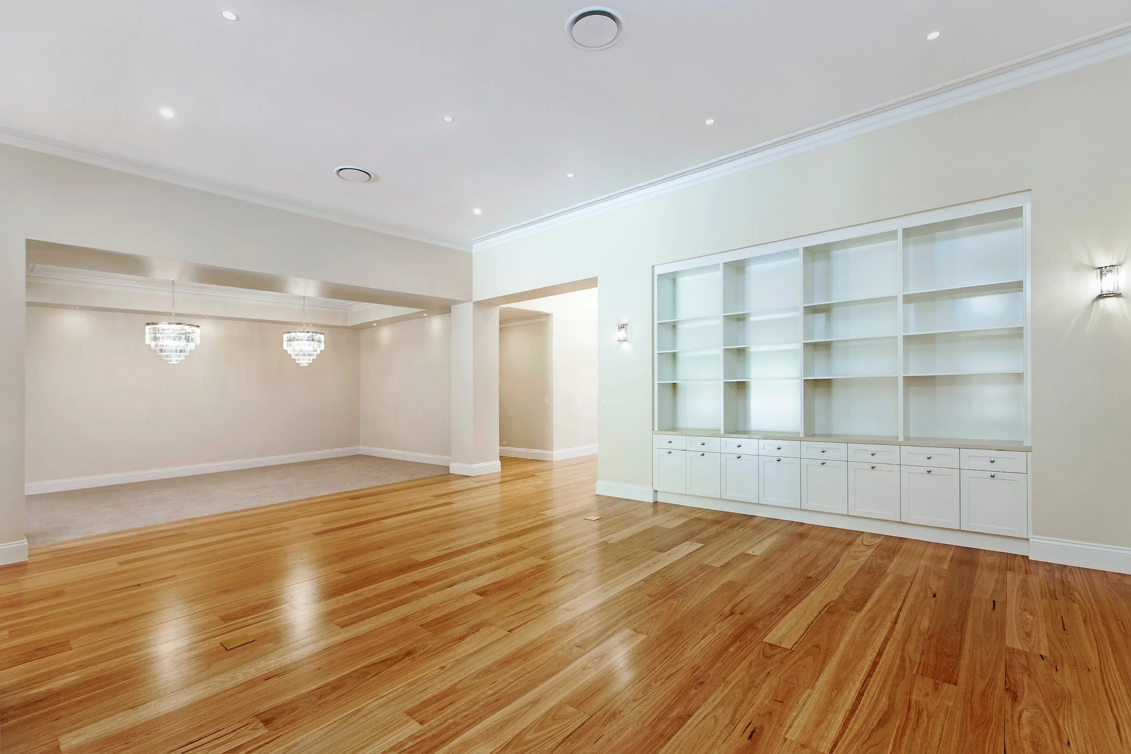 An empty living room with wooden flooring, white walls, built-in white shelving, and modern chandeliers.
