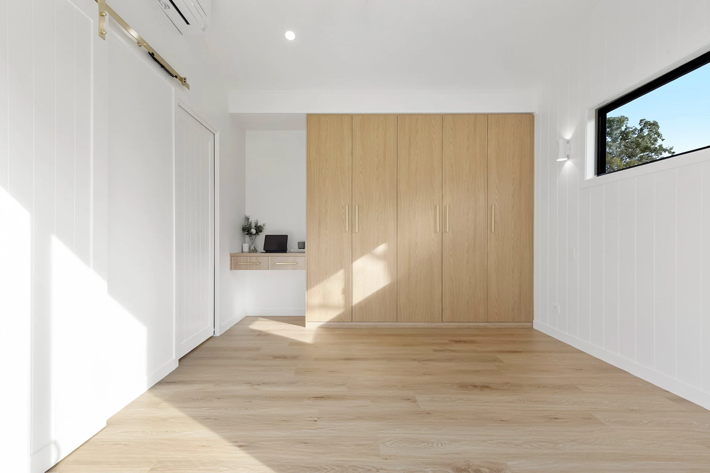 Modern interior room with light wood flooring, white walls, a large window letting in natural light, and a built-in wooden closet.
