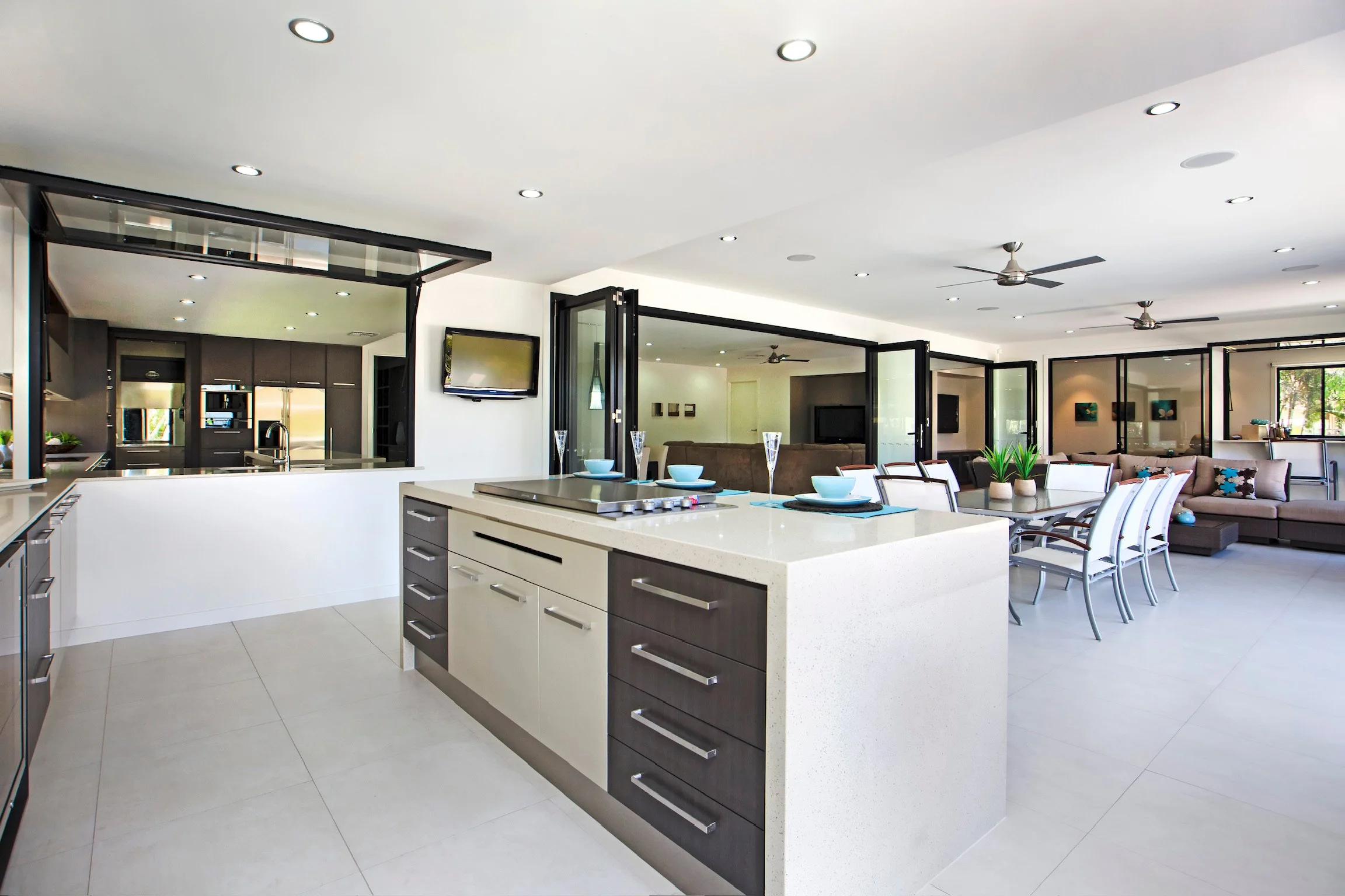 Modern open-concept kitchen and dining area with white countertops, dark wood cabinets, and a large island. The space extends into a living area with a sofa, tables, and a television. Large windows and glass doors allow natural light to fill the room