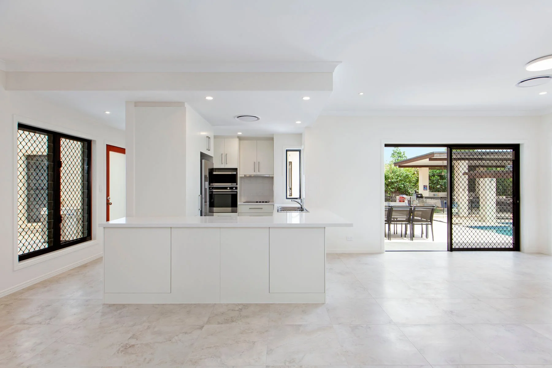 Modern white kitchen with large sliding glass doors leading to outdoor seating and pool area, granite countertop island, and natural light.