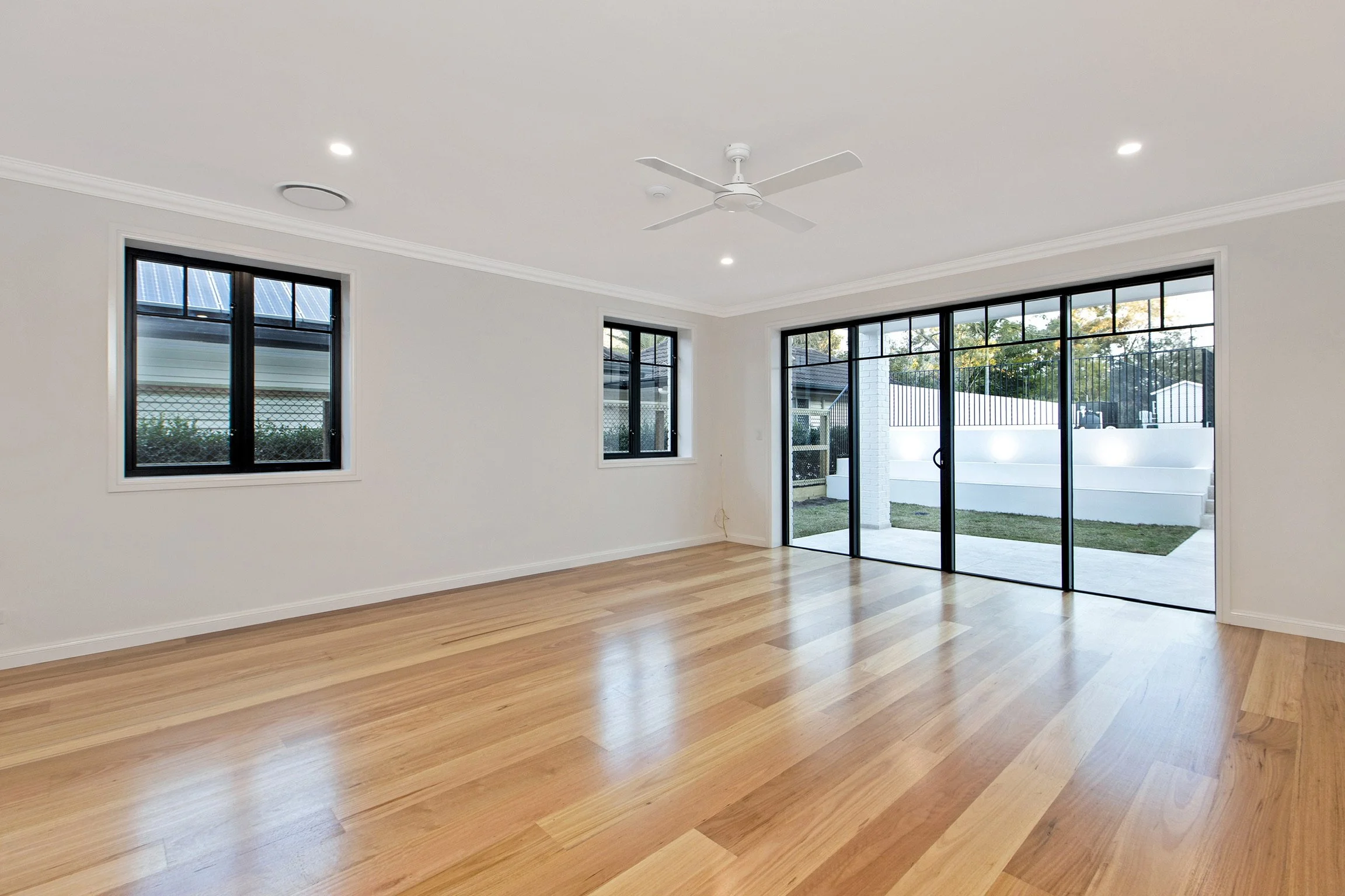 Empty living room with large sliding glass doors, two windows, hardwood flooring, white walls, ceiling fan, and recessed lighting.