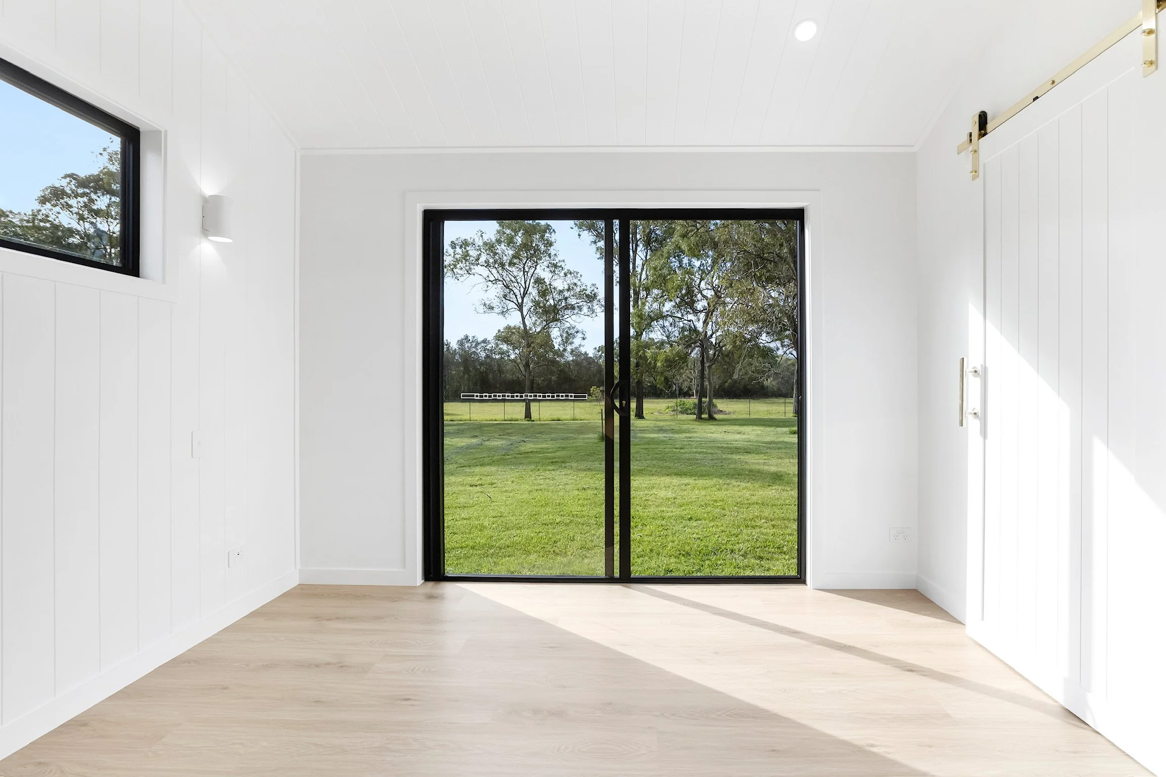 Interior view of a room with white walls, light-colored wooden flooring, and a large sliding glass door leading outside to a lawn with trees.
