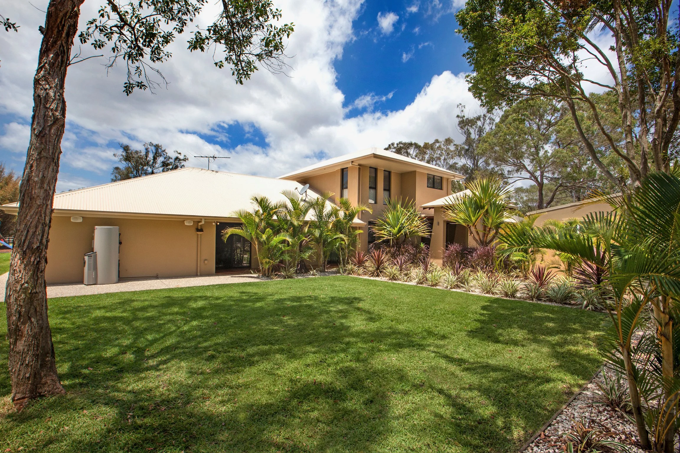 A modern house with a beige exterior, surrounded by a well-maintained lawn, tropical plants, and tall trees under a partly cloudy sky.
