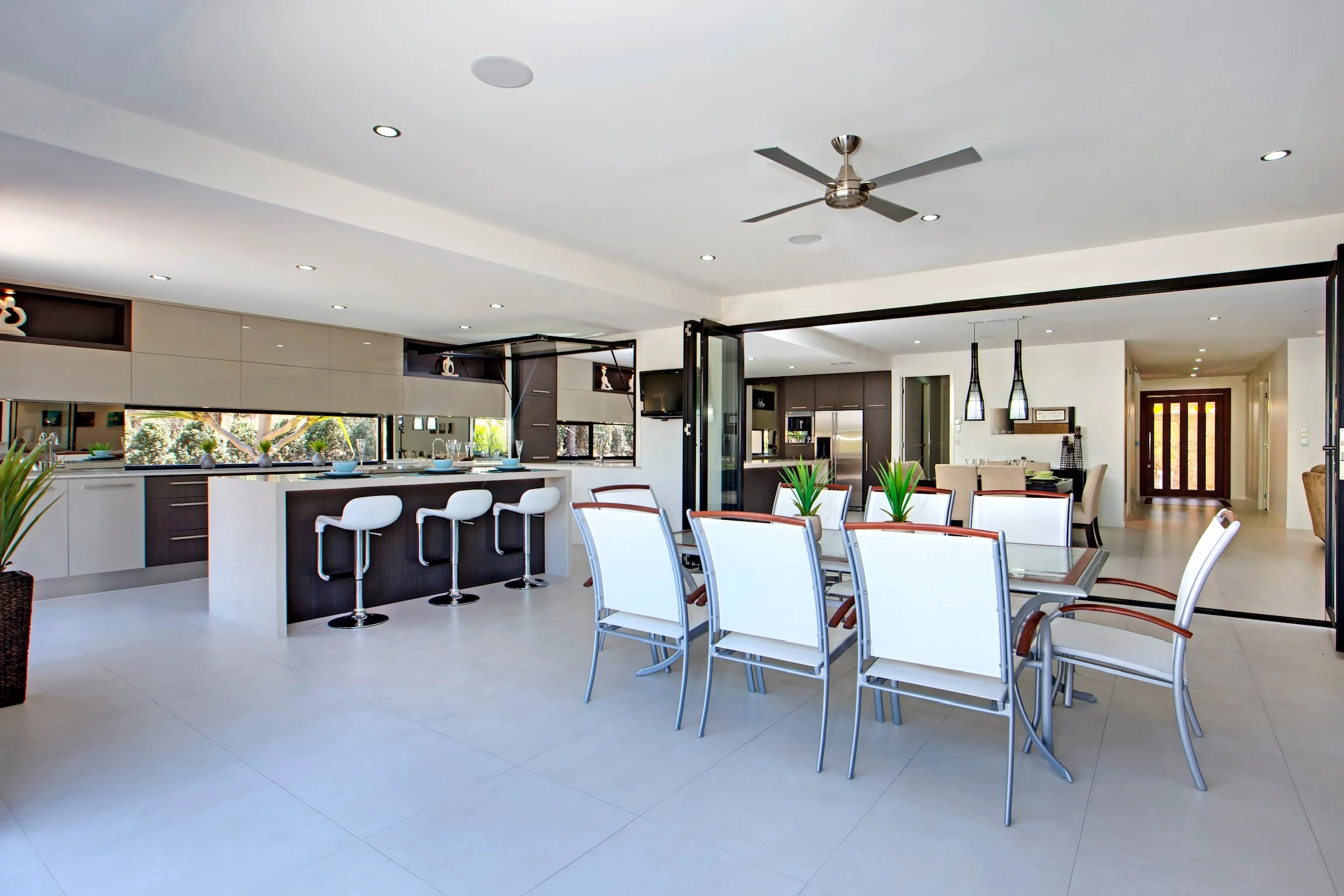 Modern open-concept kitchen and dining area with white and dark cabinetry, large windows, a ceiling fan, and multiple dining tables with white chairs and green plants.