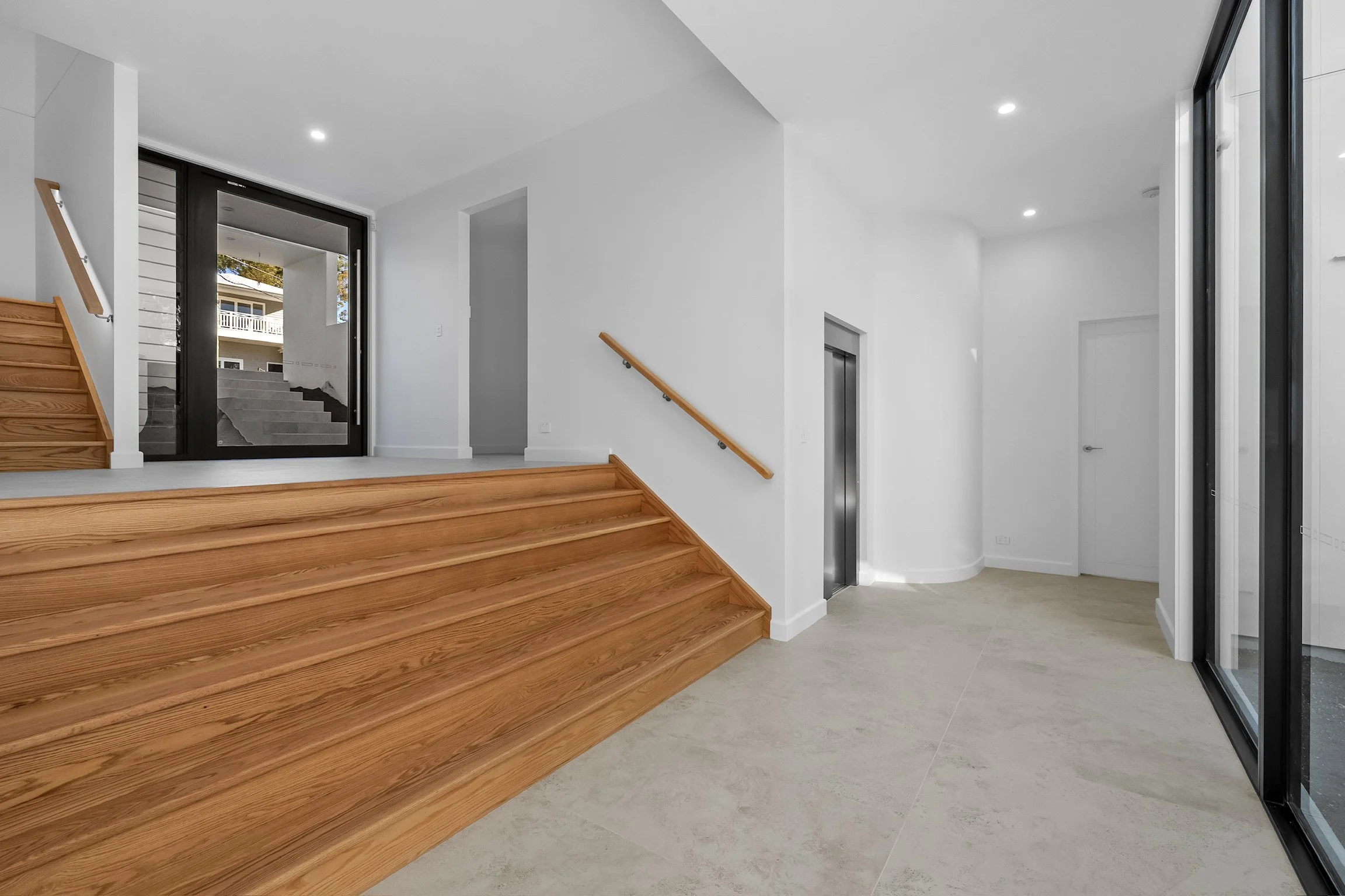 Interior of a modern house with wooden stairs, a glass door near the stairs leading outside, an elevator, and a white wall with recessed lighting.