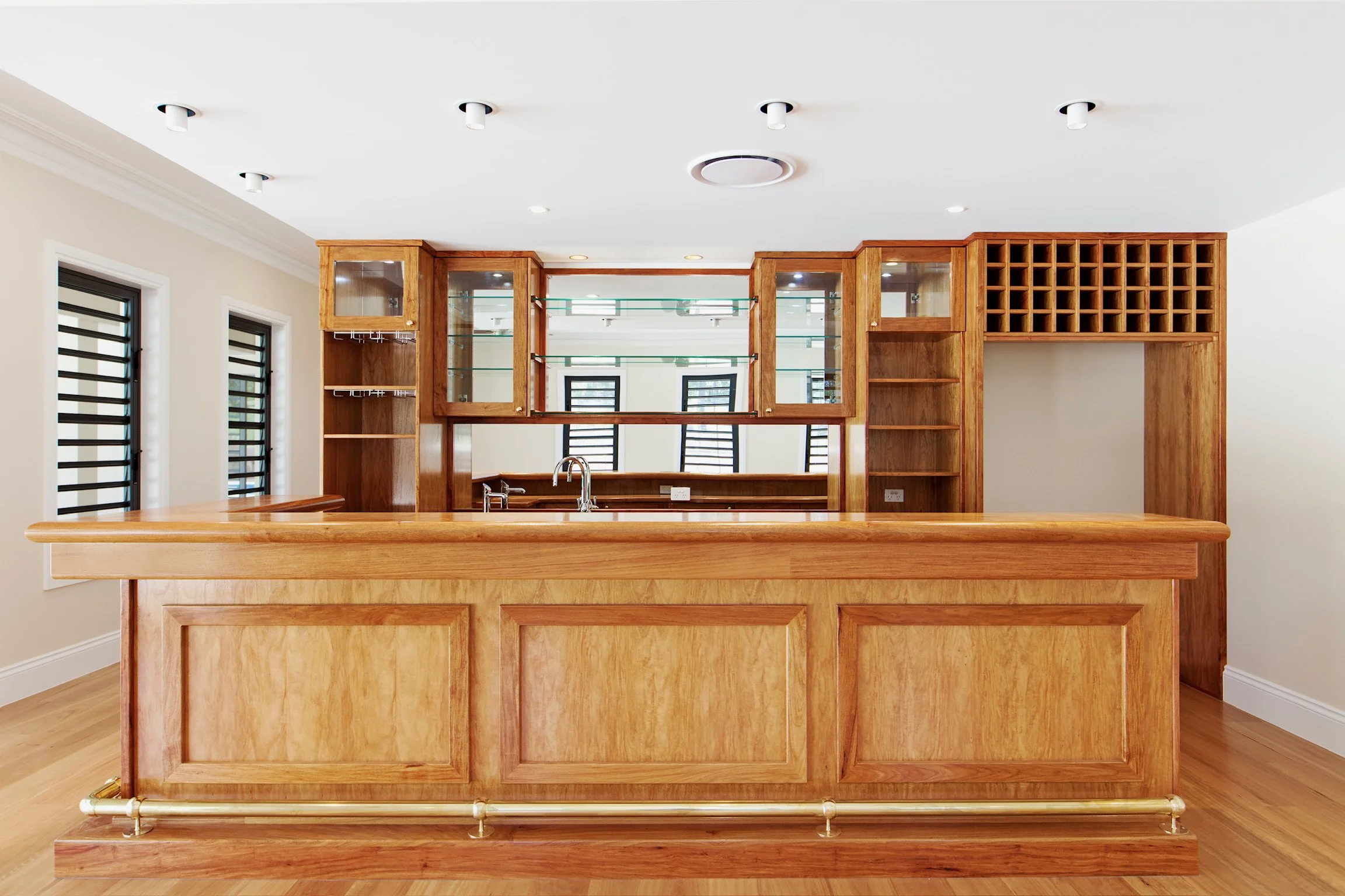 Wooden bar counter with built-in shelves and a glass rack, set in a room with white walls, windows with black shutters, and a ceiling with multiple recessed lights.