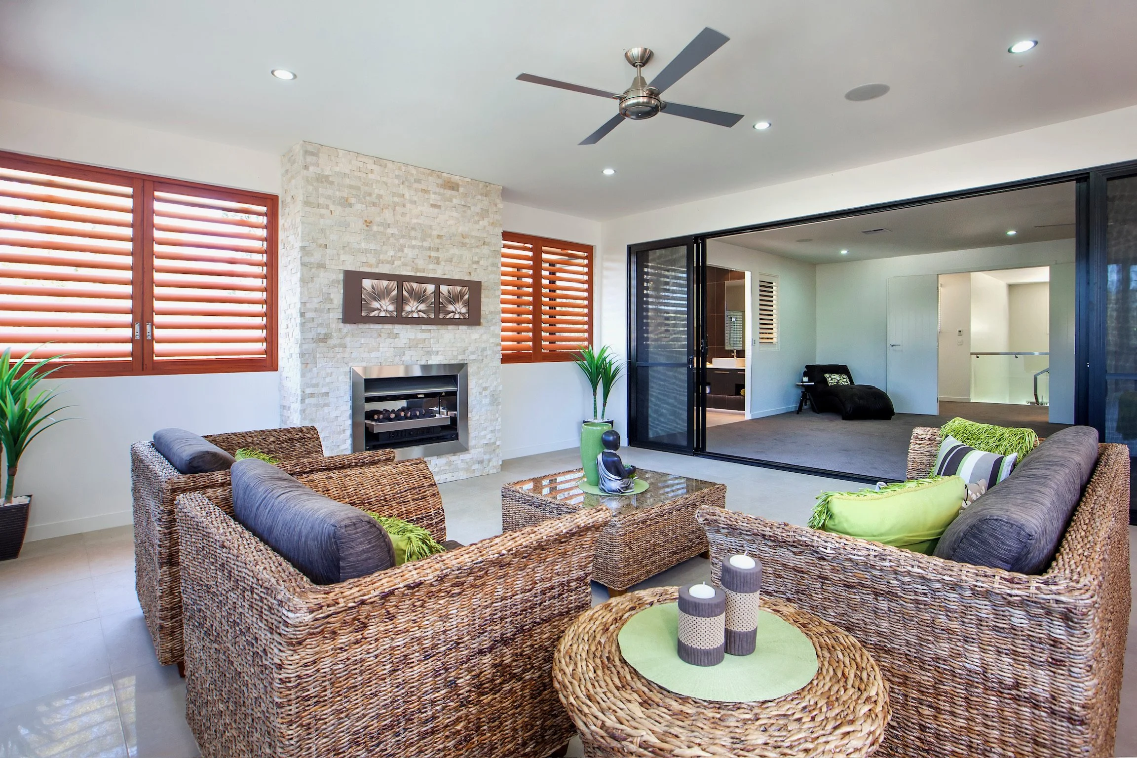A living room with wicker armchairs around a glass coffee table, a white stone fireplace with three framed art pieces above it, large windows with wooden blinds, a ceiling fan, and sliding glass doors leading to another room.