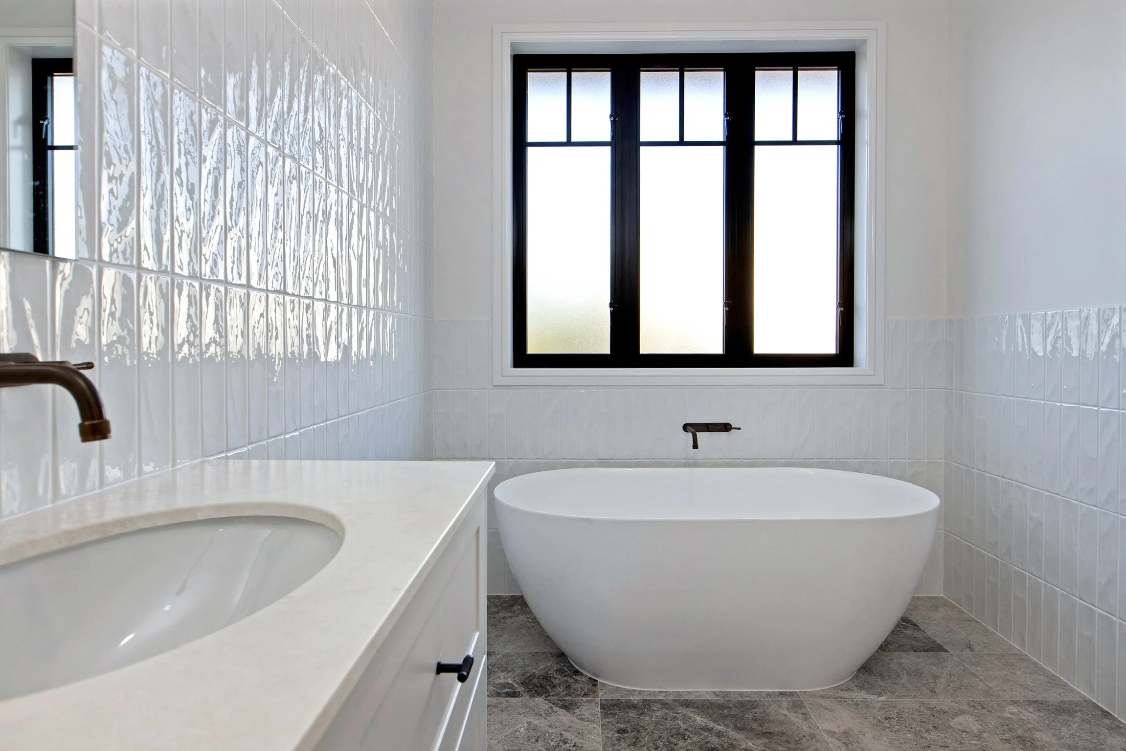A bathroom with white tiled walls, a large frosted window, a white countertop with a sink, and a large white bathtub.
