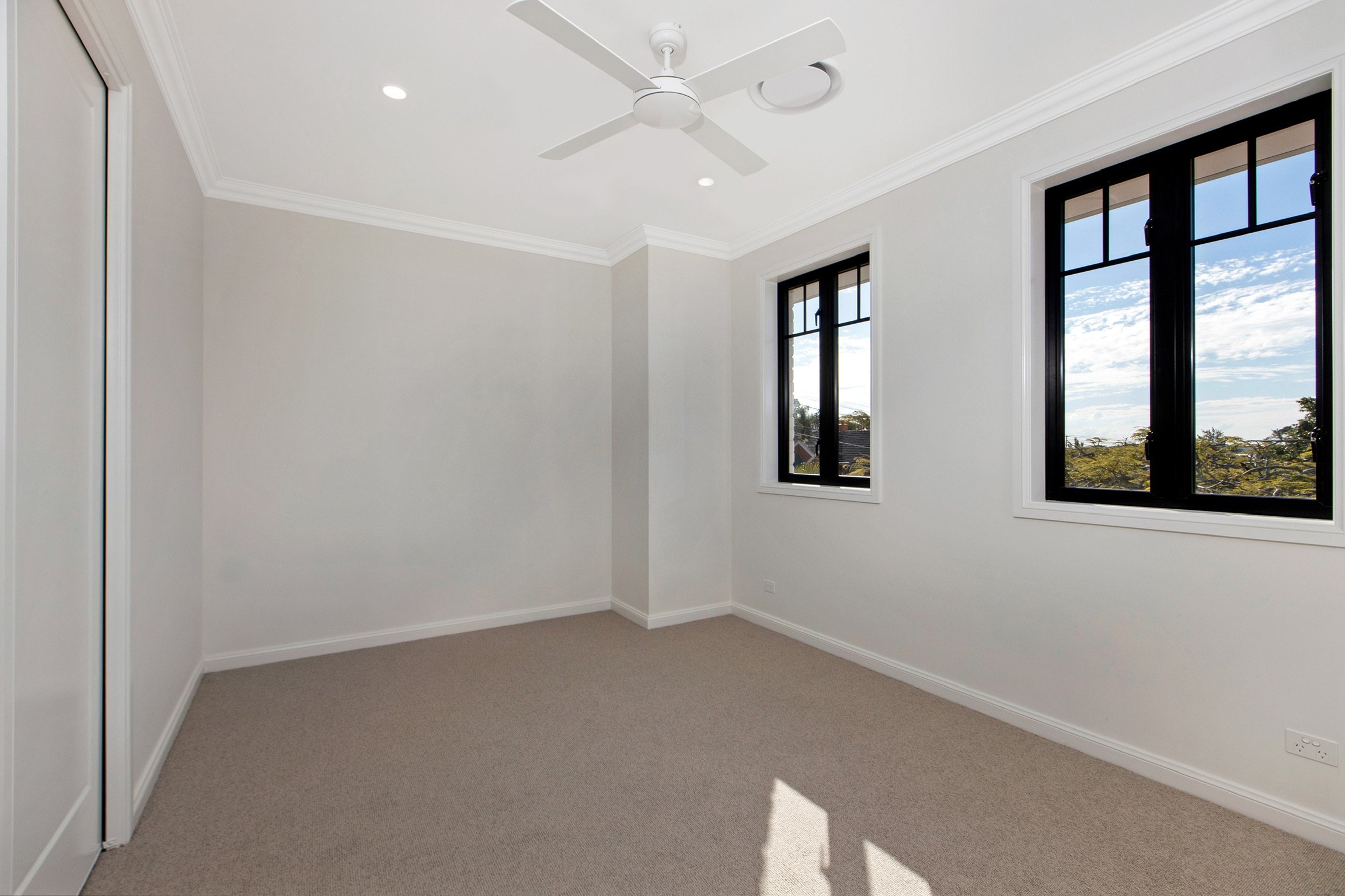 Empty bedroom with beige carpet, white walls, two black framed windows showing a view of trees and sky, white ceiling fan, and a sliding door with mirror.
