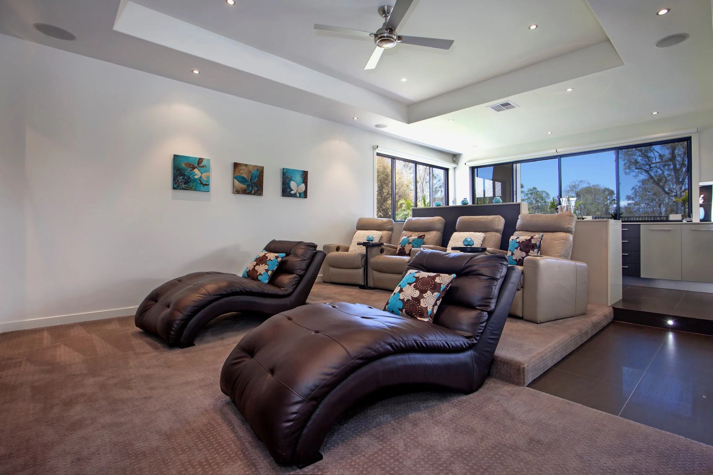Living room with leather recliners and beige sofa, decorated with butterfly-themed pillows, large windows with trees outside, and artwork of butterflies on the white wall.