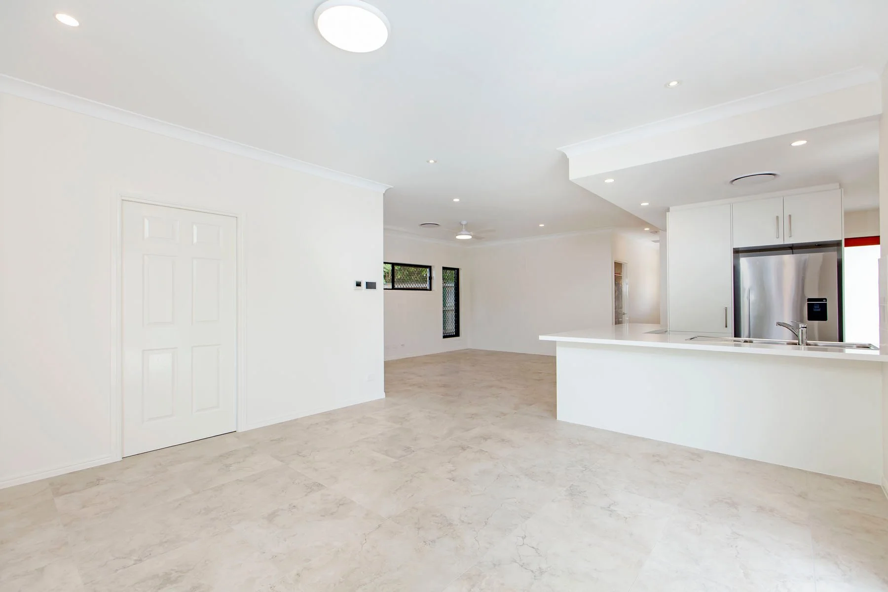 Empty living room and kitchen area with white walls, beige marble flooring, stainless steel refrigerator, white cabinets, and recessed ceiling lights.
