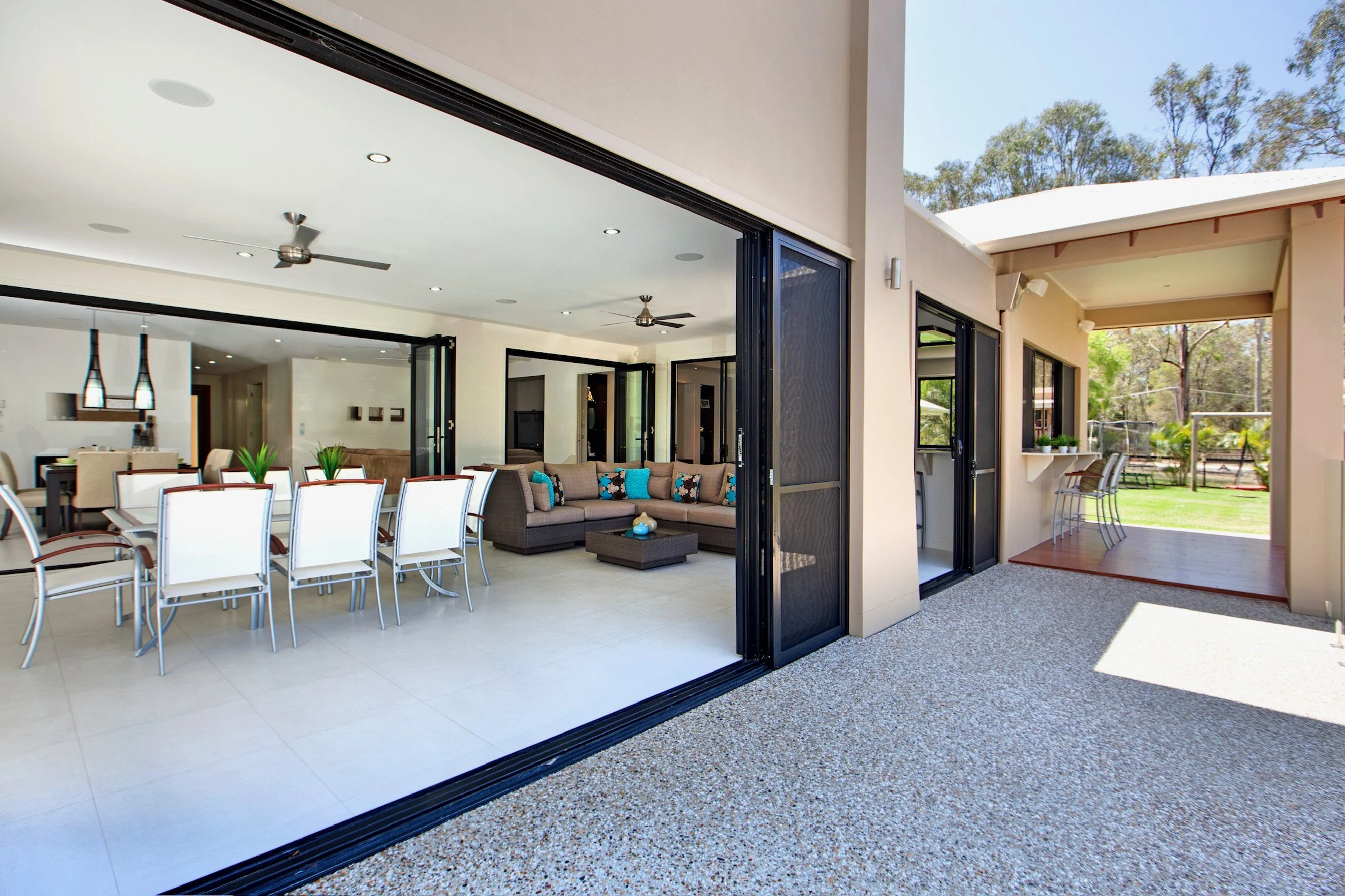 Open sliding glass doors leading from a bright indoor dining area with white chairs and a living room to an outdoor patio with chairs and greenery.