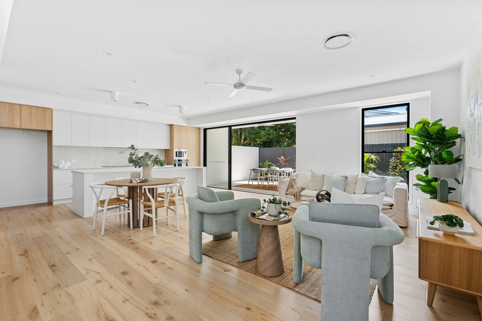 Spacious, modern living room and kitchen area with wooden floors, white cabinetry, a dining table, and large windows opening to an outdoor patio with greenery.