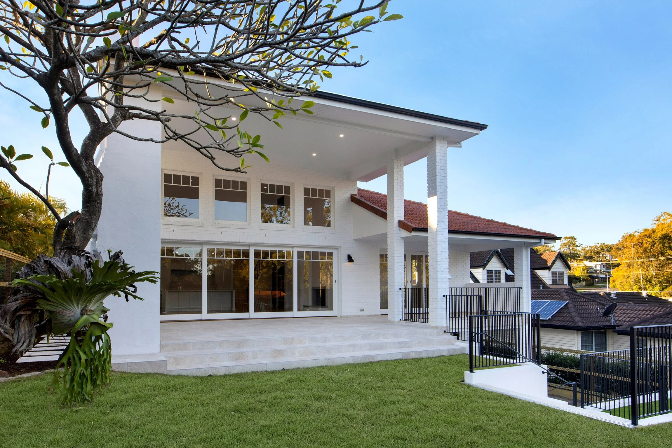A modern white house with large glass sliding doors and a balcony with a black metal railing. The house has a sloped roof, and the yard features green grass, trees, and plants, with a clear blue sky in the background.