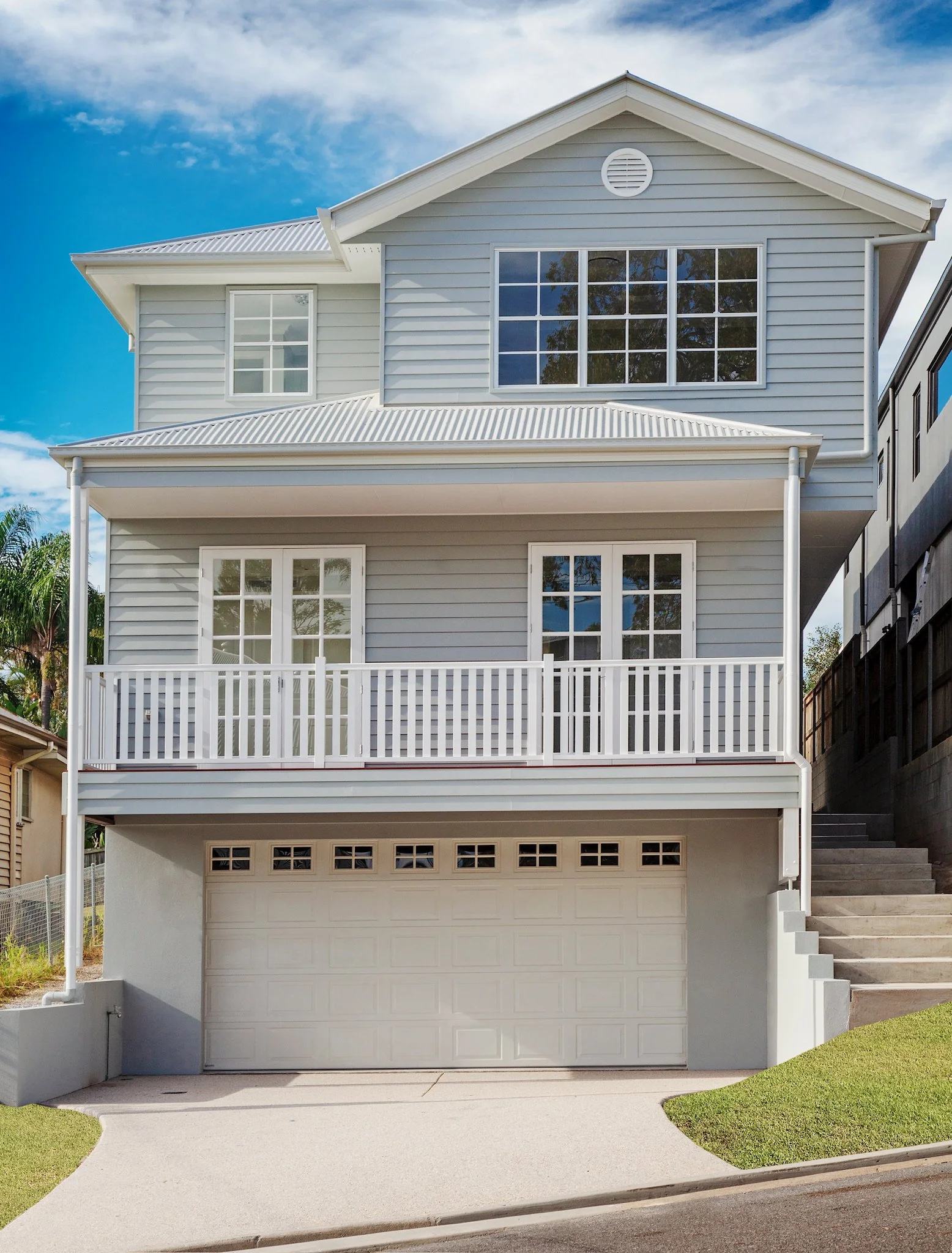 A modern three-story house with a white garage door, a balcony with a white railing, large windows, and a grey exterior with horizontal siding.