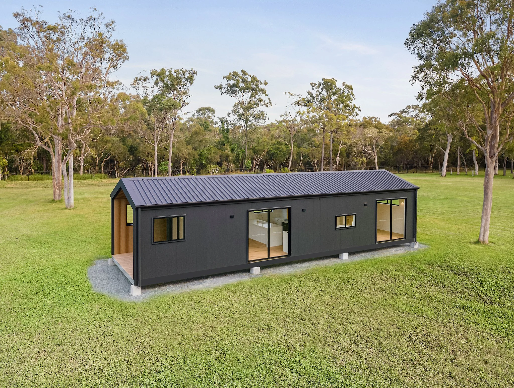 A small modern house with a black exterior and large glass windows is situated on a green lawn, surrounded by trees and a fence, under a clear sky.
