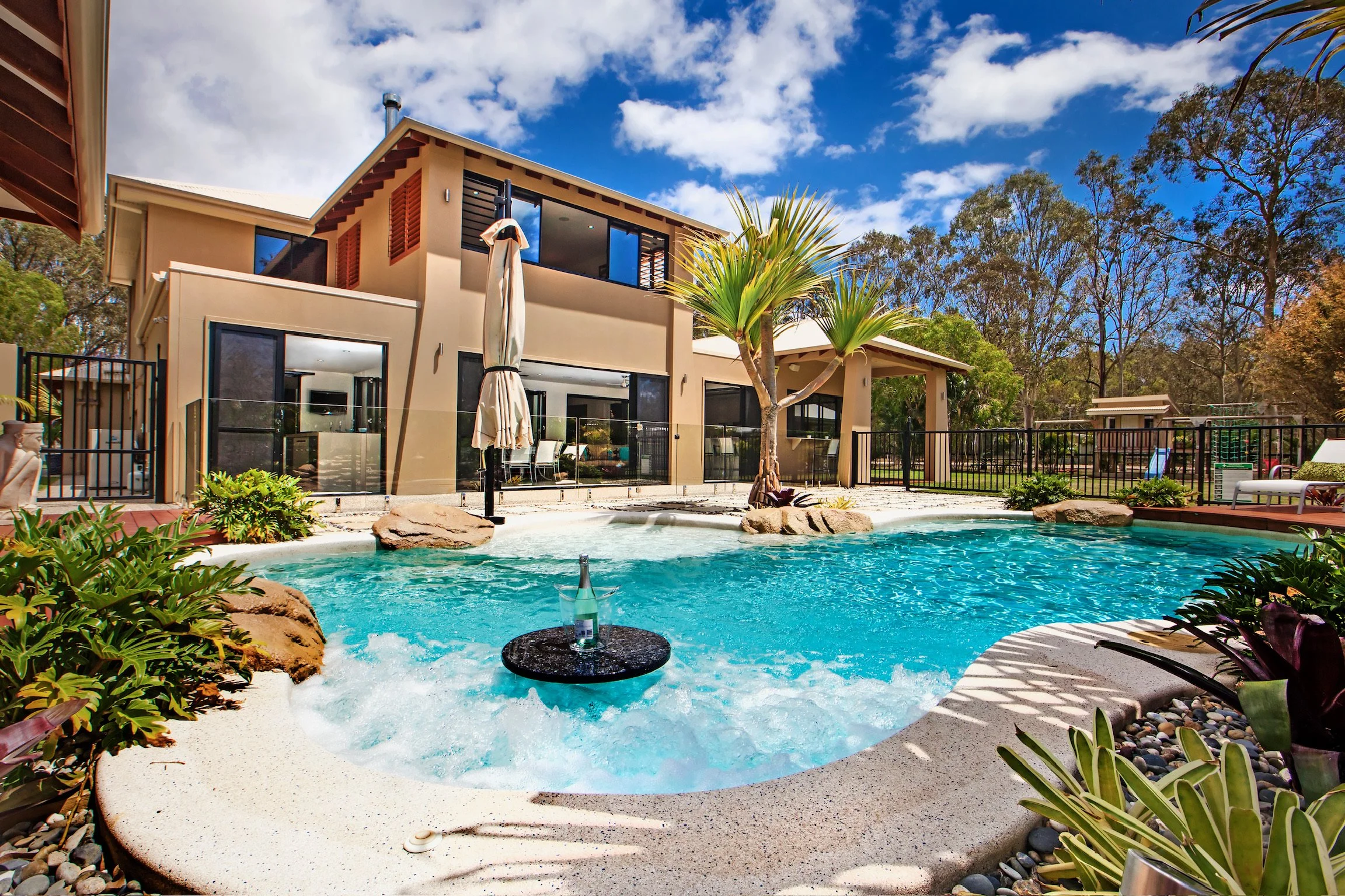 A backyard with a swimming pool, surrounded by plants and trees, with a two-story house in the background. The house has large windows and a balcony on the upper level. The sky is blue with some clouds.