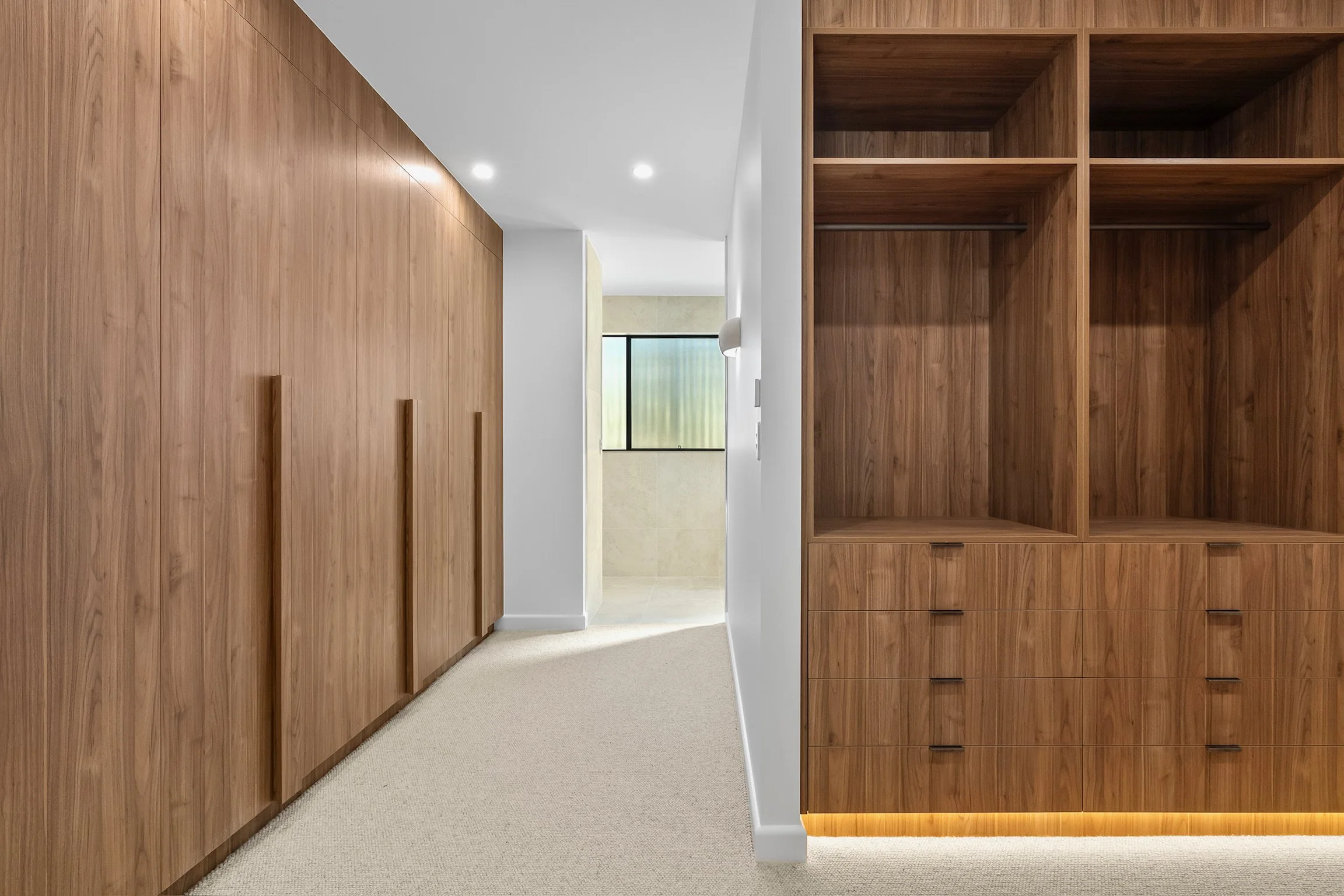 A hallway with wooden cabinets on the right and a beige carpeted floor, leading to a room with a window in the distance.