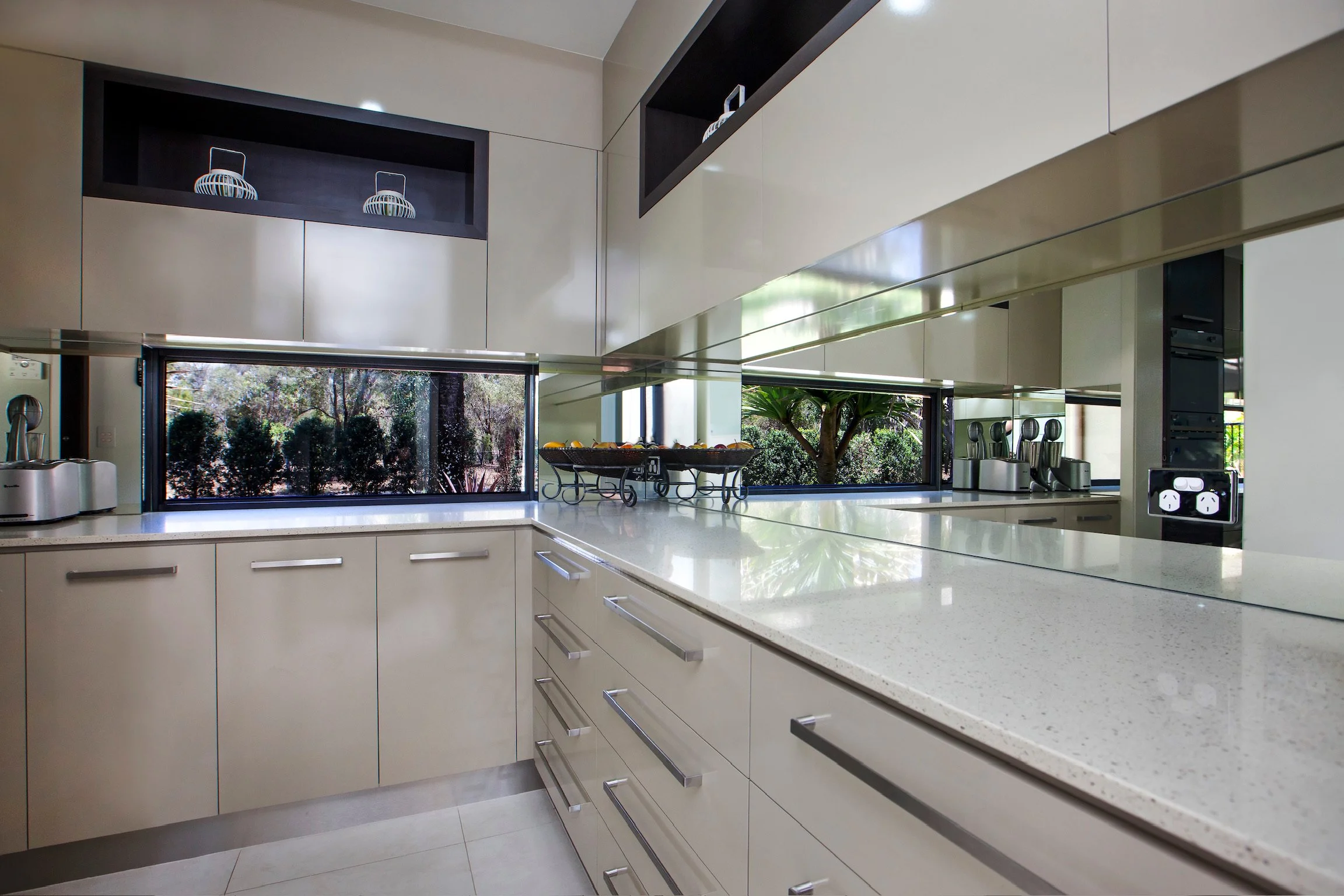 Modern kitchen with beige cabinets, white speckled countertops, large windows with trees outside, and stainless steel appliances.