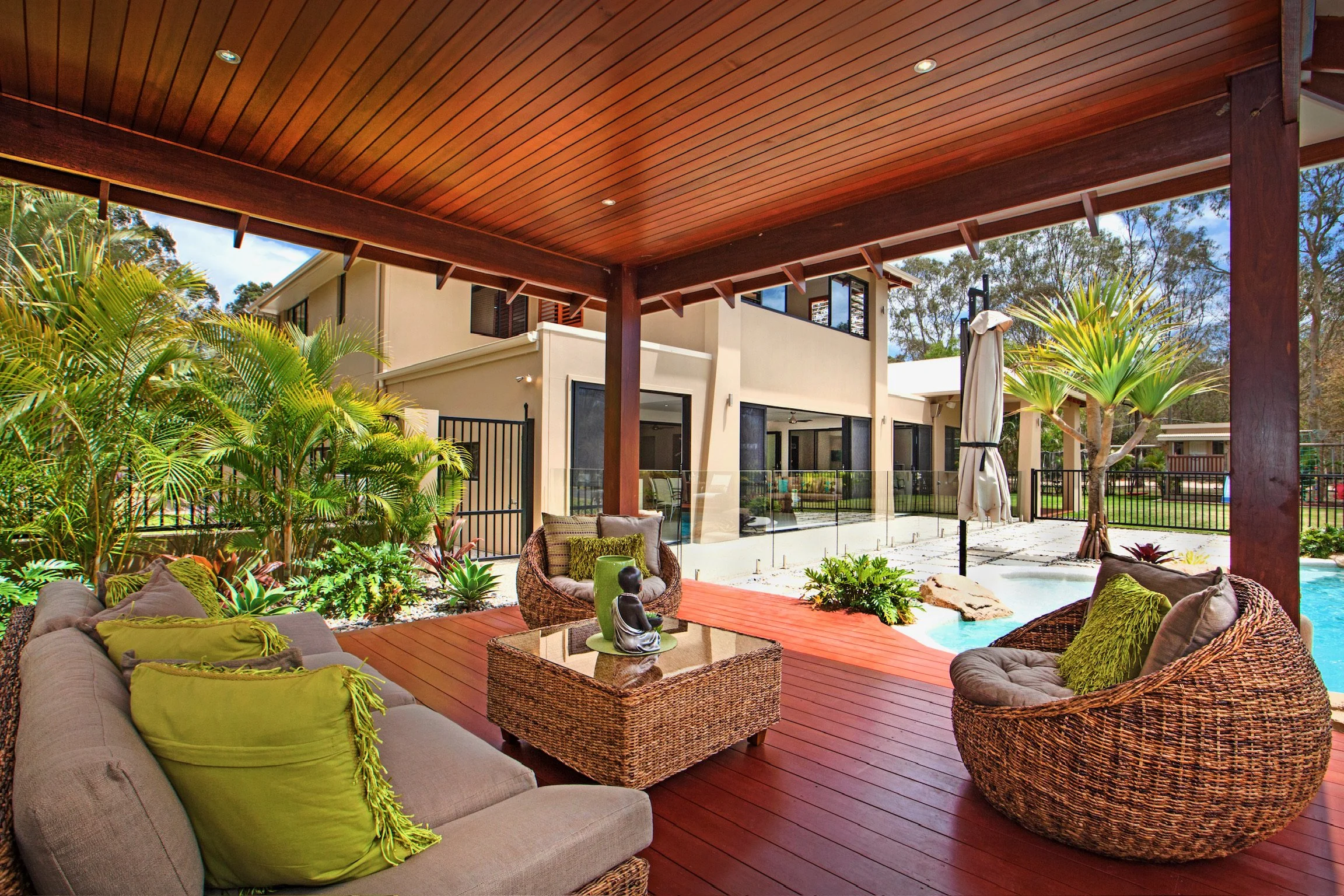 Outdoor patio with wicker furniture and green pillows under a wooden ceiling, lush plants, and a modern house in the background.