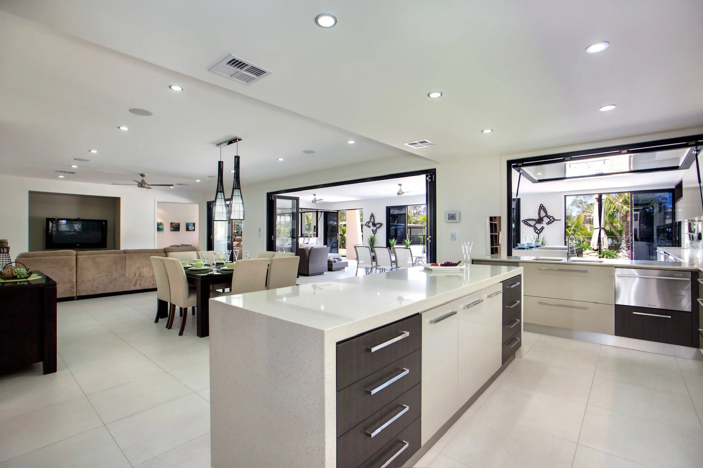 Open-concept dining and living area with white walls, tiled flooring, a kitchen island with dark drawers and a white countertop, and large open windows showing outdoor greenery.