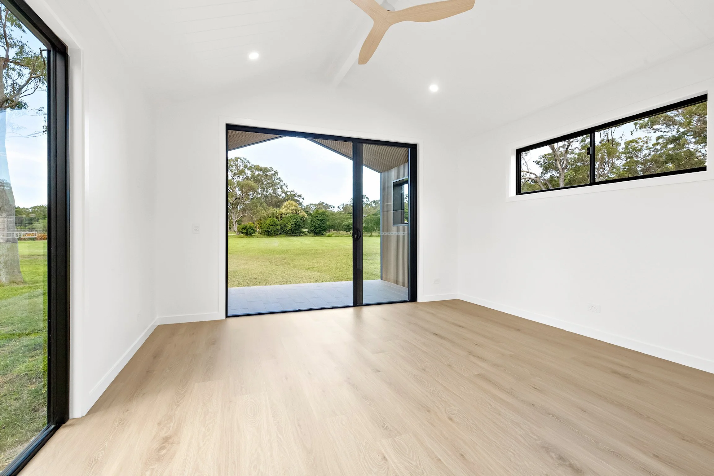 Empty room with large sliding glass doors and windows, showing a green yard and trees outside.