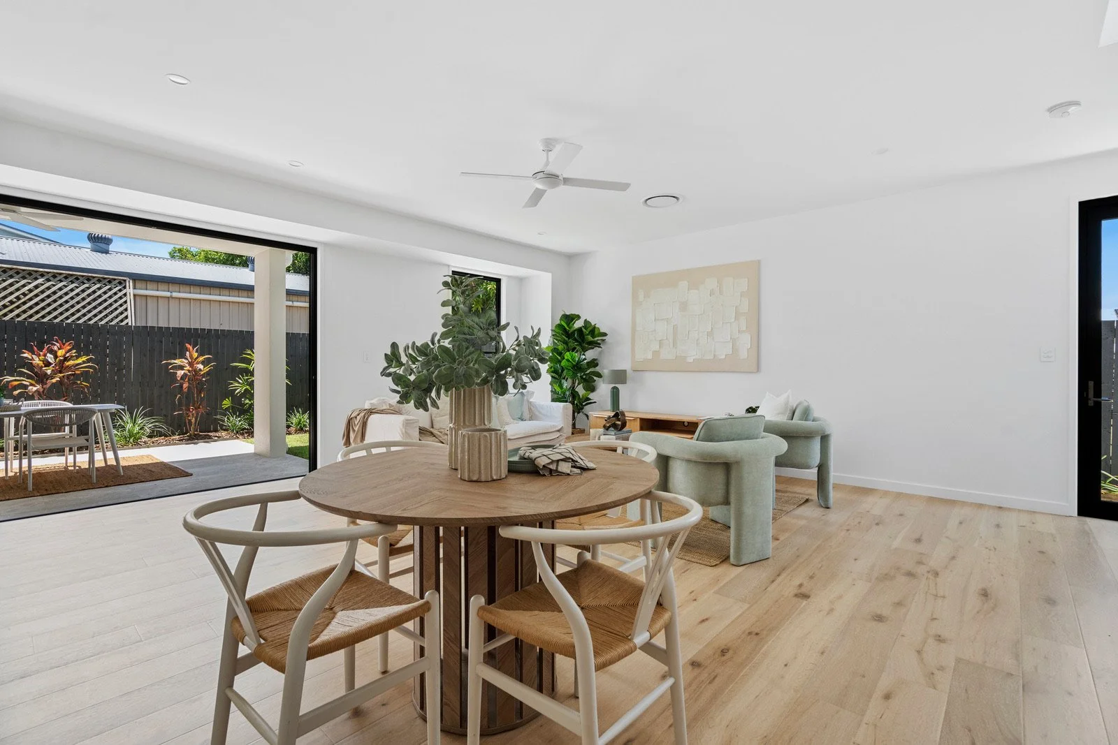 Modern living room with a dining table, chairs, a sitting area, and large sliding glass doors leading to an outdoor patio with garden plants.