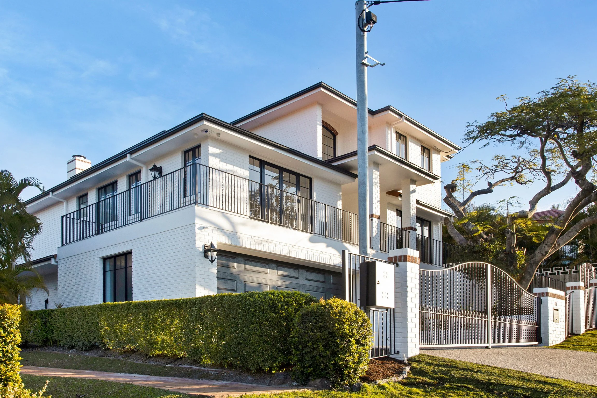 A white multi-story house with black trim, balconies, and a gated fence, surrounded by greenery and a blue sky.