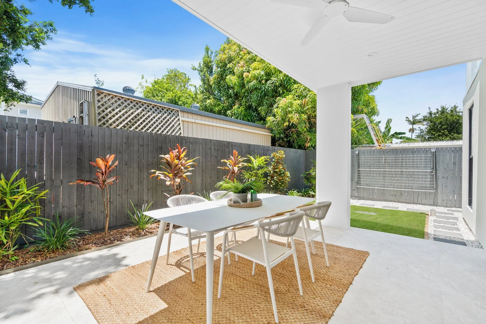Outdoor patio area with white table and chairs, plants, and a woven rug, enclosed by a wooden fence, under a ceiling fan.