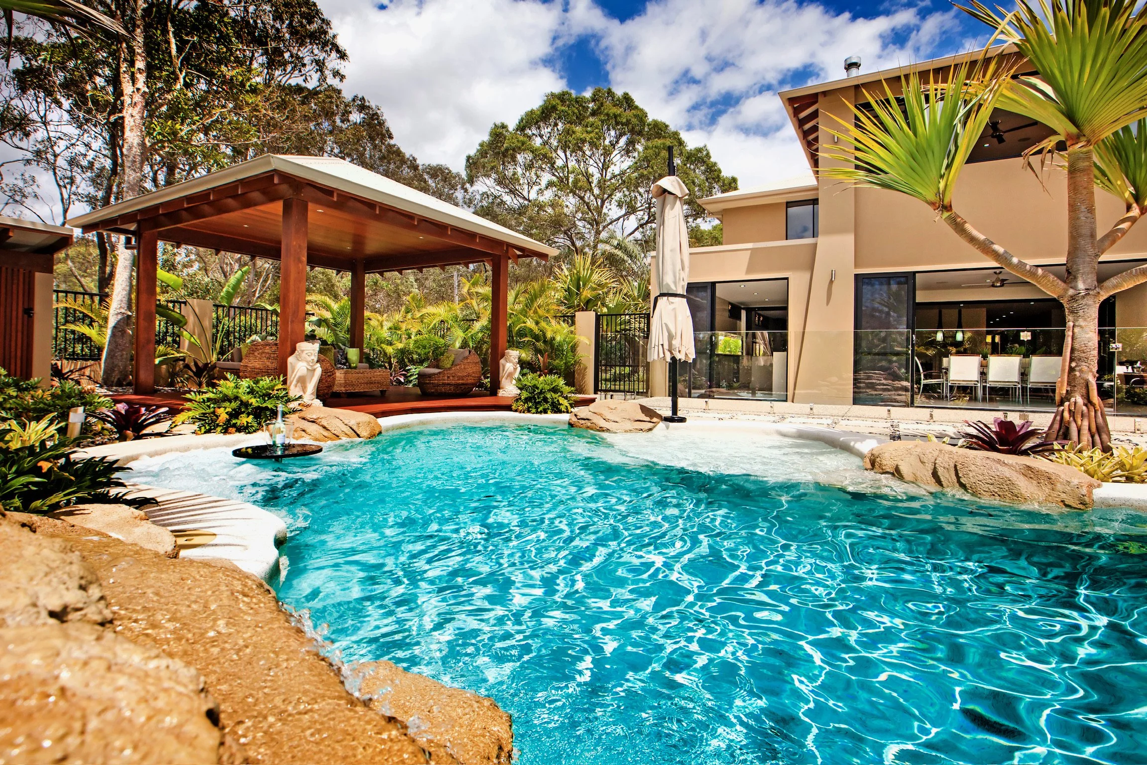 An outdoor swimming pool with clear blue water, surrounded by plants, rocks, and a modern house with a patio and lounge area.