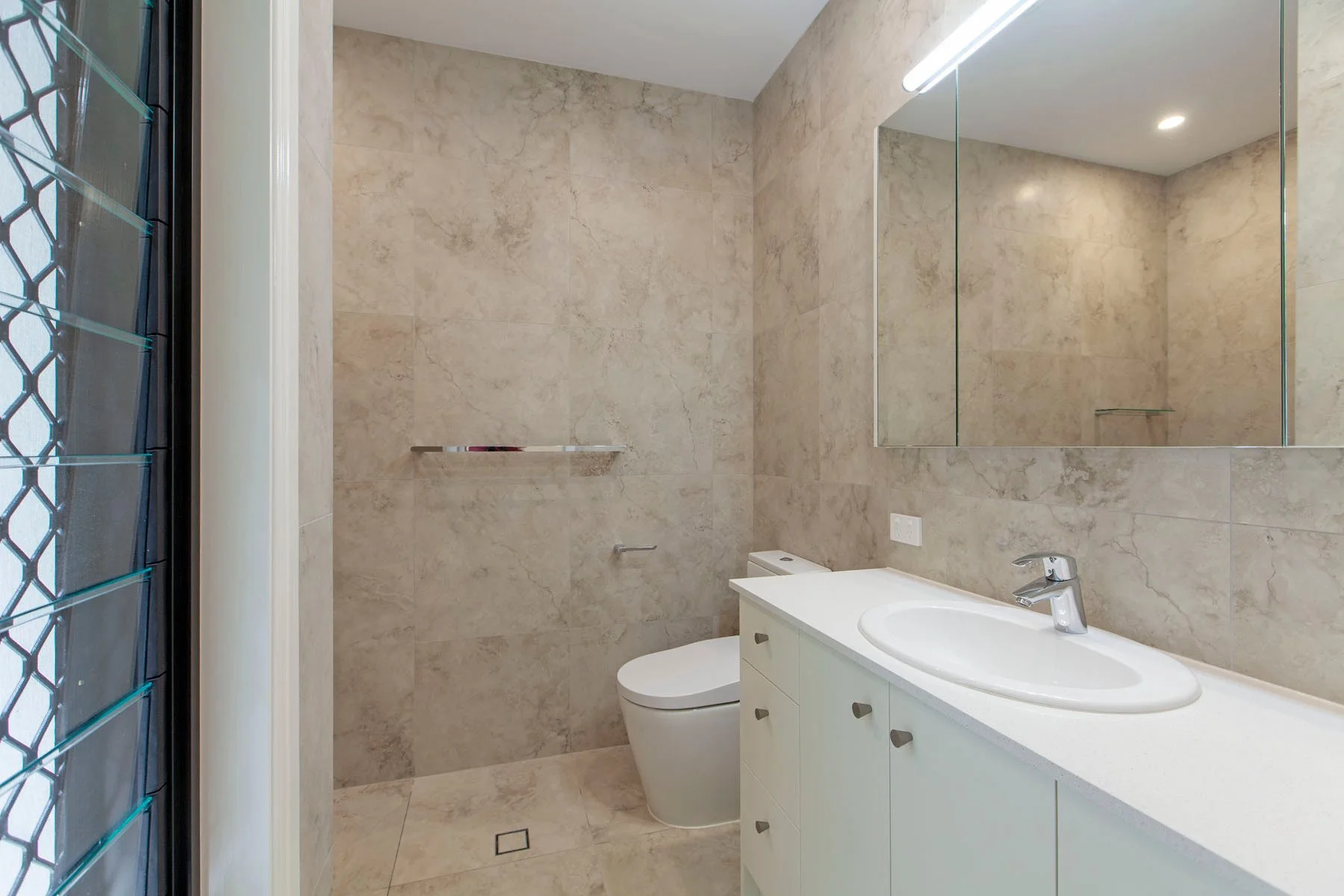 A modern bathroom with beige marble tiles on the walls and floor. There is a white vanity with a sink and a chrome faucet. A large rectangular mirror is above the vanity. To the left, there is a window with black security bars. The bathroom includes 