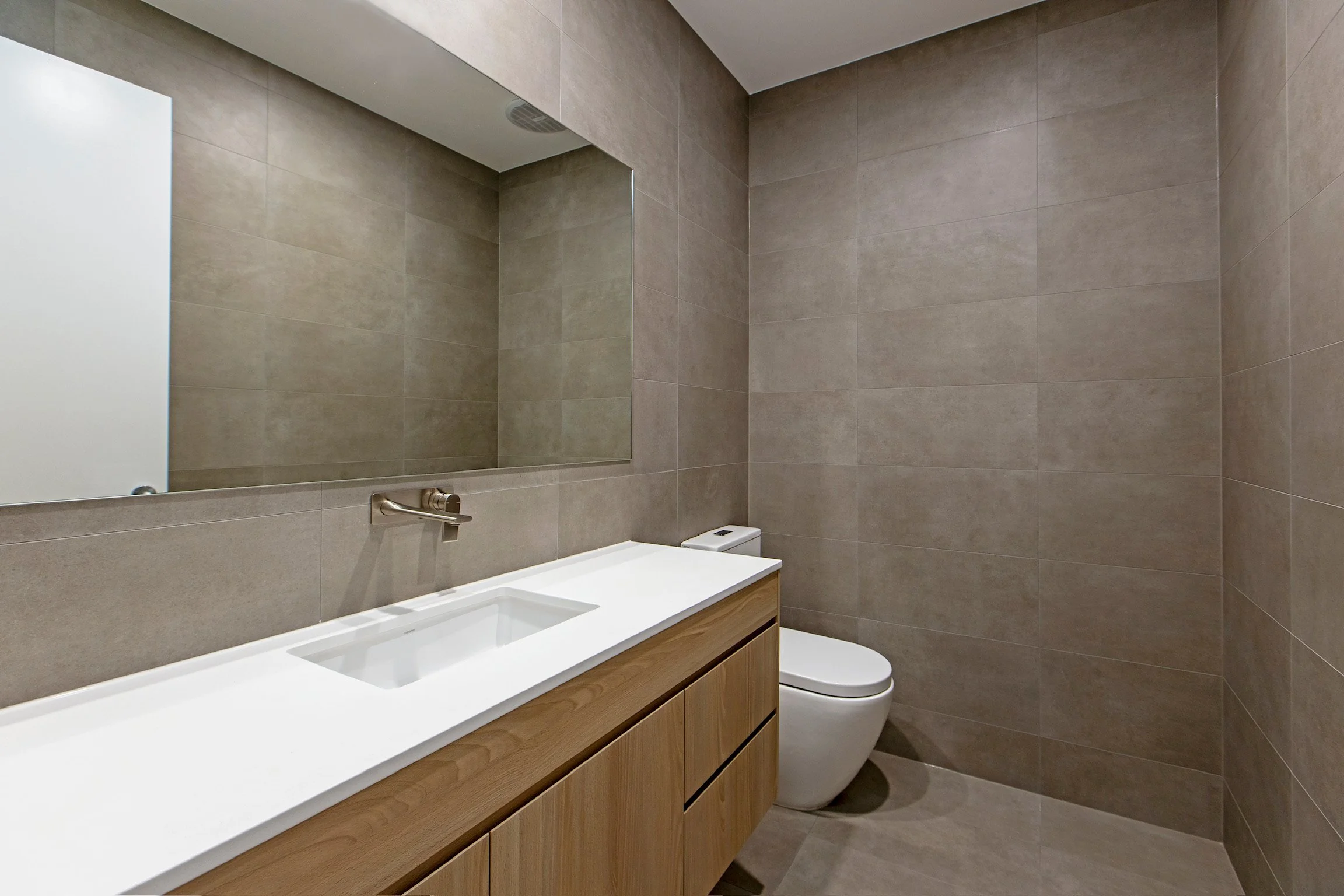 A modern bathroom with beige tiled walls, a large mirror, a white sink with a wooden cabinet below, a toilet, and a ceiling vent.