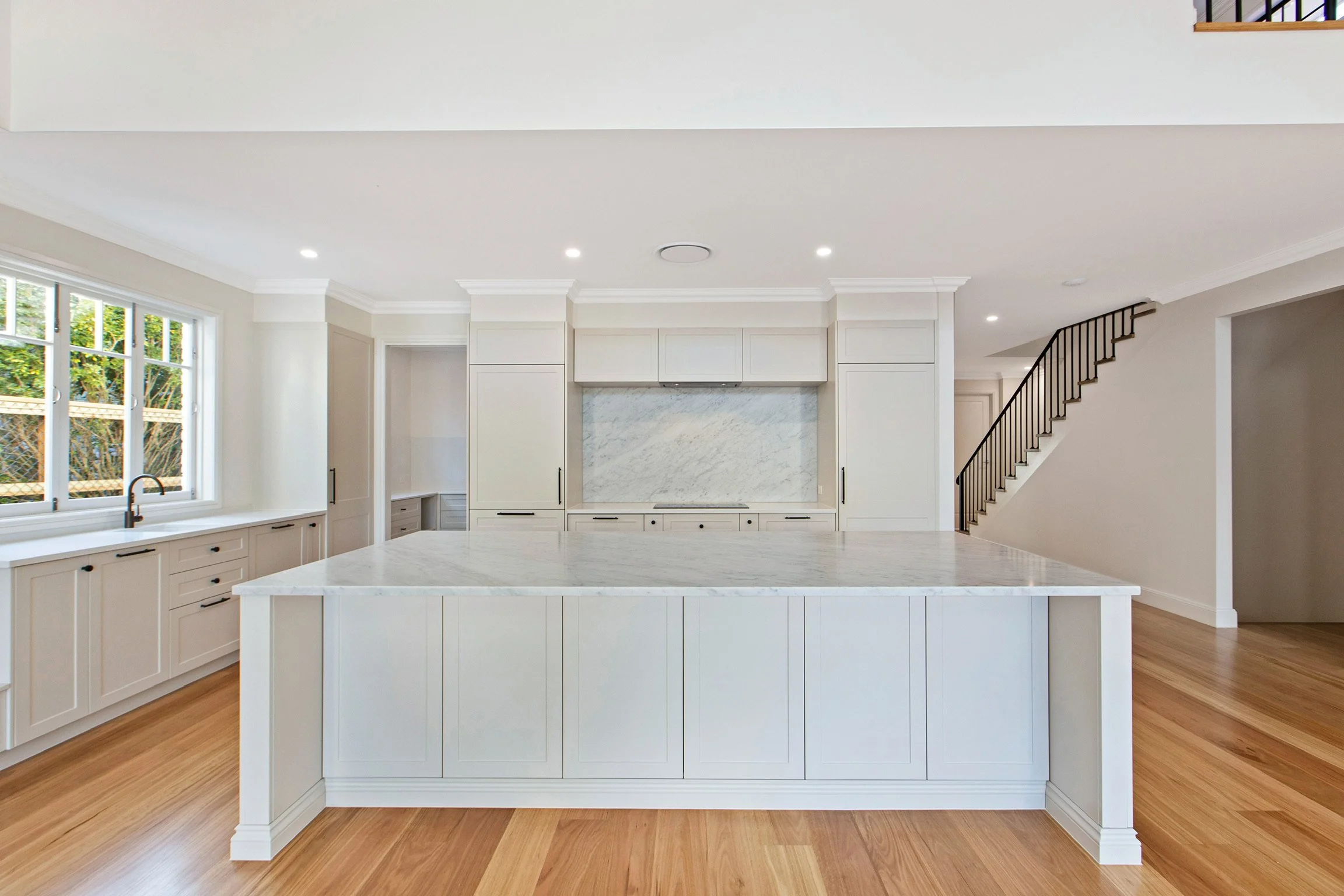 Modern kitchen with white cabinetry, marble island, large window, and staircase with black railing.
