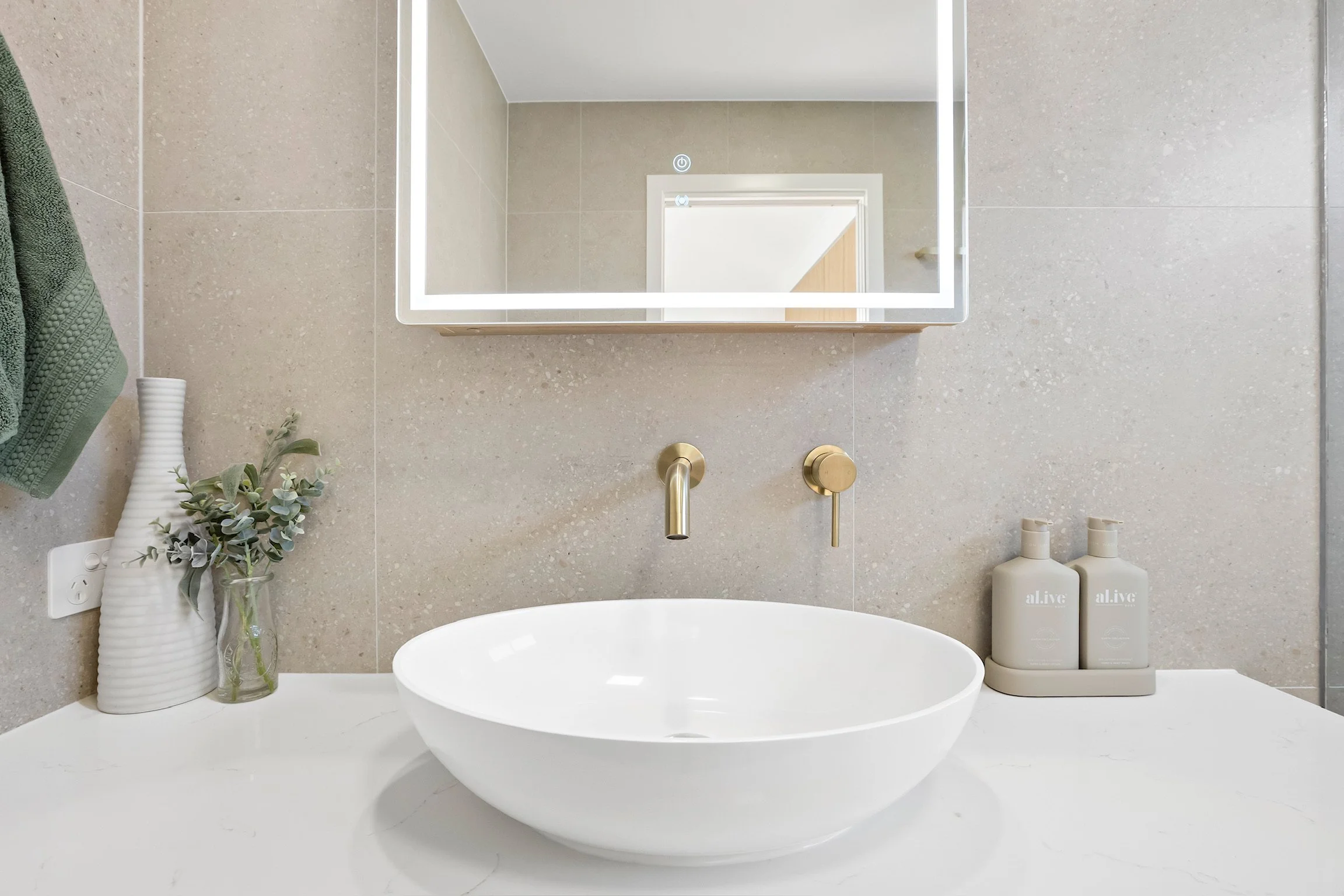 A modern bathroom mirror above a white sink, with a beige tiled wall. Decorative items include a white vase with green foliage and a glass container with more greenery. Two beige bottles labeled 'alive' are beside the sink.