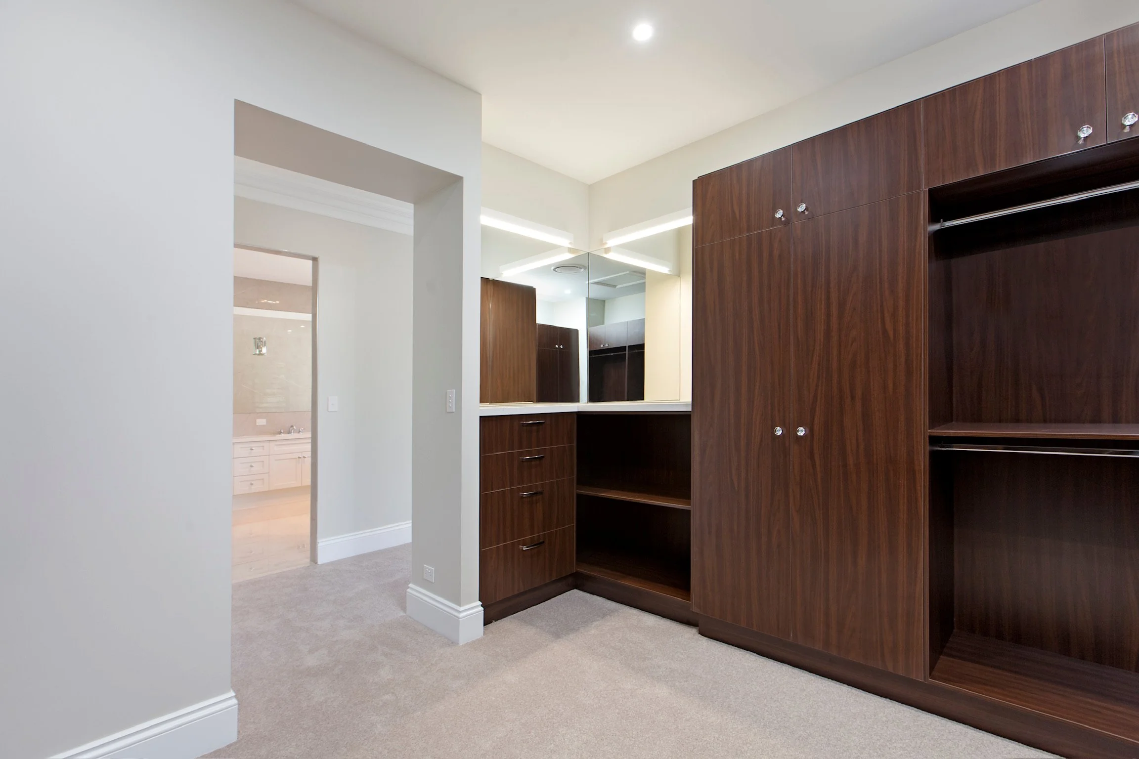 Room with beige carpet, white walls, built-in wooden closet, overhanging mirror, and kitchen area visible in the background.