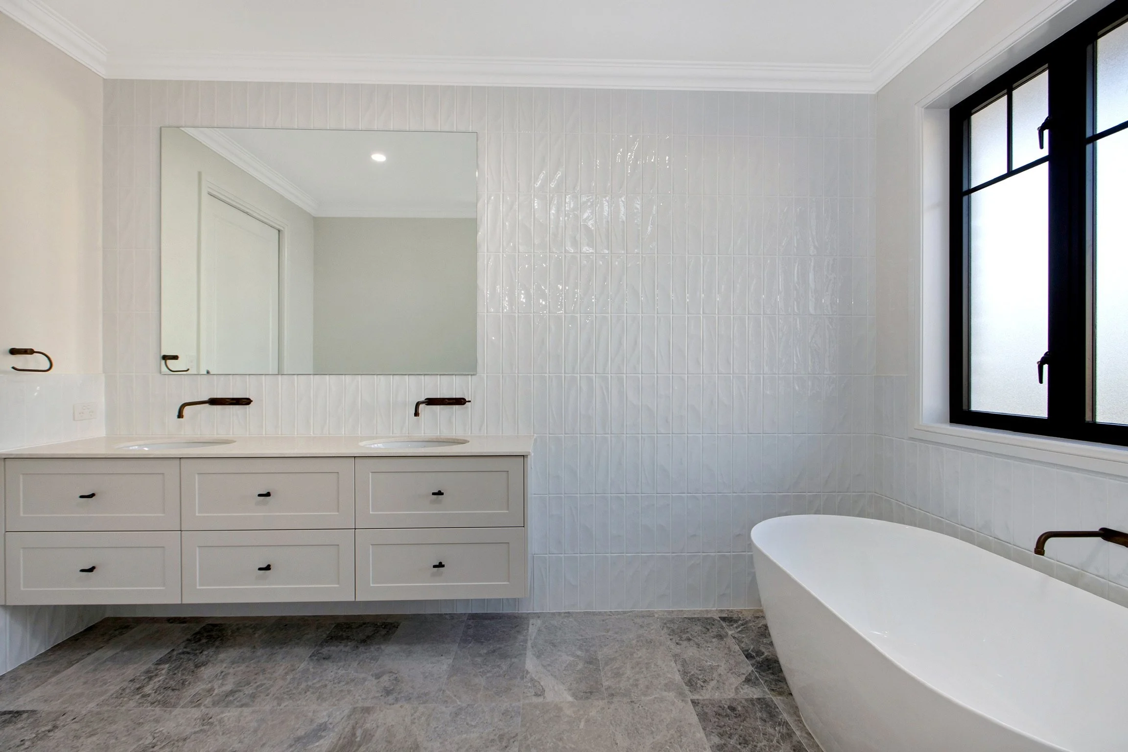 Modern bathroom with gray tile flooring, white vanity with two sinks, large mirror, black fixtures, frosted window, and a white bathtub.