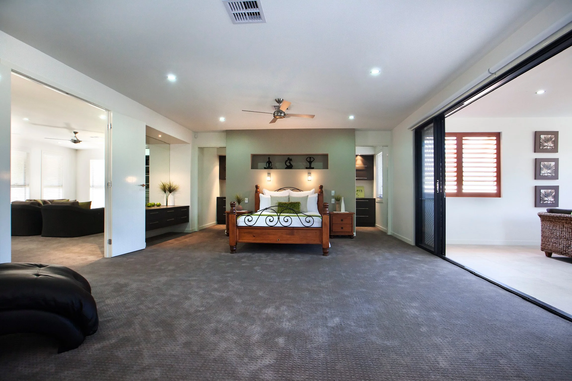 Interior of a spacious bedroom with a wooden bed, nightstands, decorative shelves, and modern lighting. An adjacent sitting area with black chairs, large windows, and a sliding glass door leading outside.