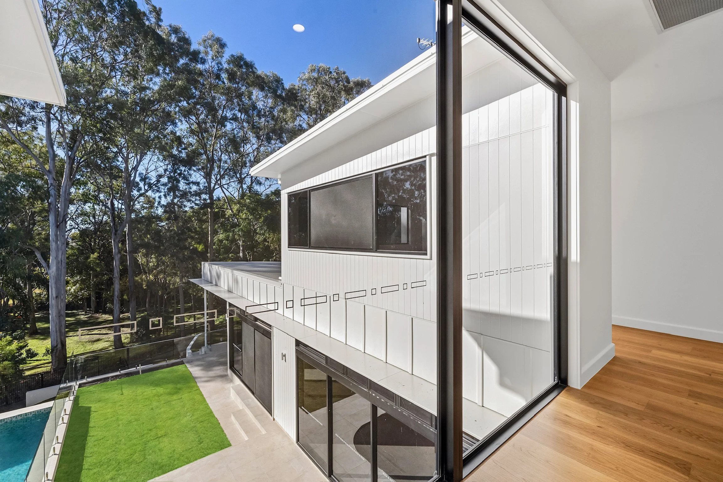 Modern house with white exterior, large windows, and a deck overlooking a backyard with a swimming pool, green grass, and tall trees under a clear blue sky.