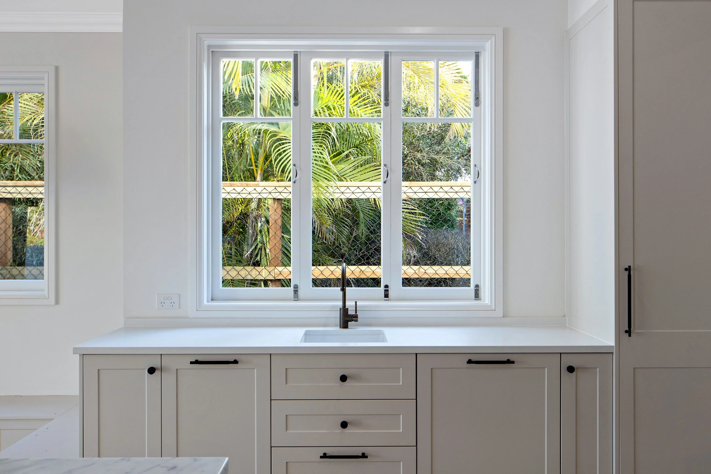 A kitchen scene with a white countertop, white cabinets, a black faucet, and a large window showing green tropical plants outside.